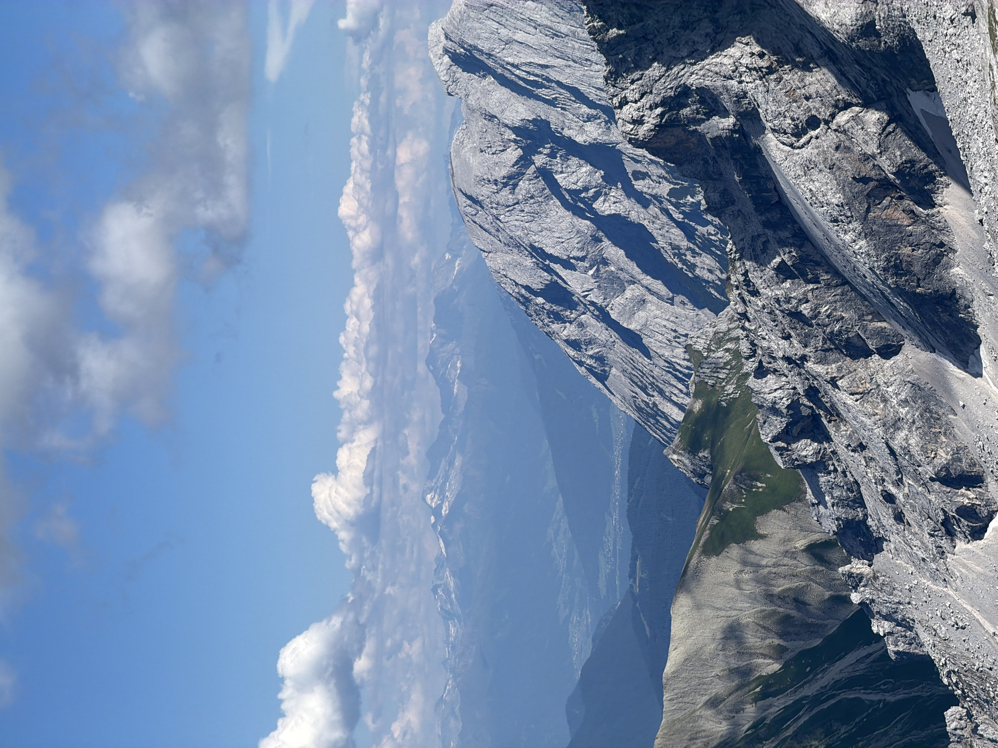 The image shows a panoramic view of a mountainous landscape under a bright blue sky with scattered white clouds. The foreground is dominated by rugged, rocky mountain slopes with visible textures of grey stone and some patches of green vegetation. In the mid-ground, layers of mountains recede into the distance, with the colors becoming progressively bluer and hazier due to atmospheric perspective. A valley with some signs of human settlement, possibly a town or village, is visible in the far distance. The overall impression is one of a vast, natural alpine environment. There are no people or specific objects clearly identifiable that would indicate a specific location within Garmisch-Partenkirchen, Germany, though the scene is consistent with the mountainous terrain surrounding the area.