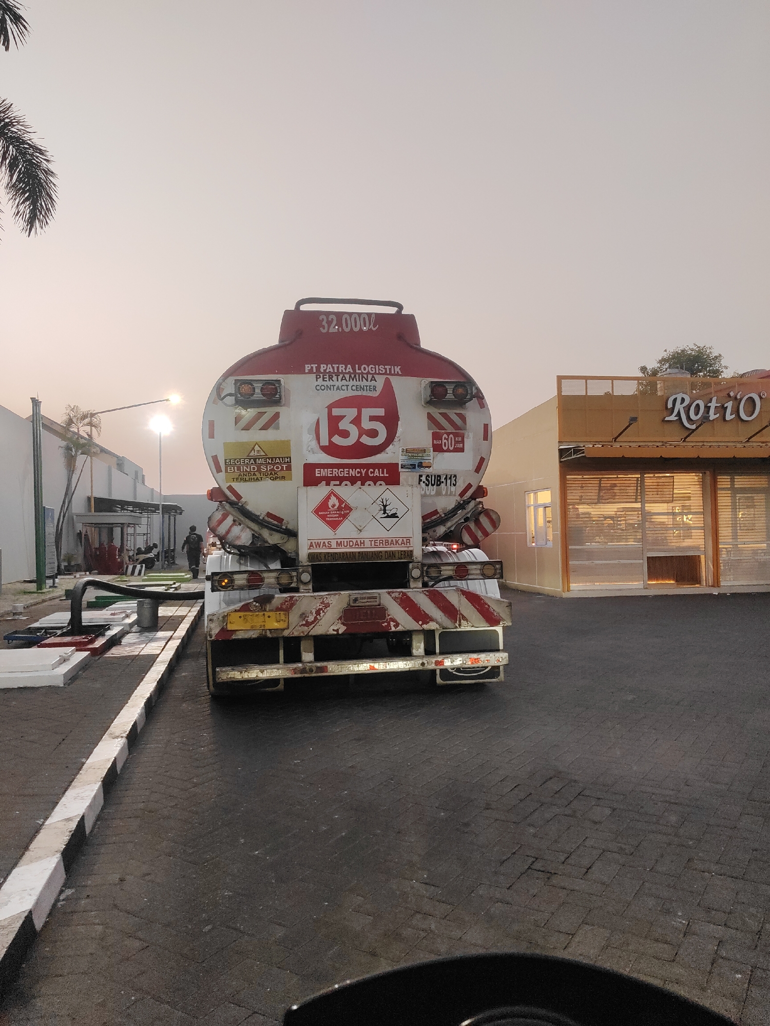 In Sidoarjo, Indonesia, at approximately 6:00 AM, a tanker truck with "PT PATRA LOGISTIK PERTAMINA CONTACT CENTER" and "32.000L" printed on its side is parked near a "Roti O" bakery. The truck's rear, marked with safety warnings and hazard symbols, faces the camera. The bakery, with its doors rolled up, reveals a warm interior. The sun, low in the sky, casts a gentle light on the scene. A street lamp, palm trees, and a building with scaffolding are visible in the background. A few individuals are seen near the scaffolding. The ground is paved with interlocking bricks, and a curb with alternating white and black stripes runs along the foreground.