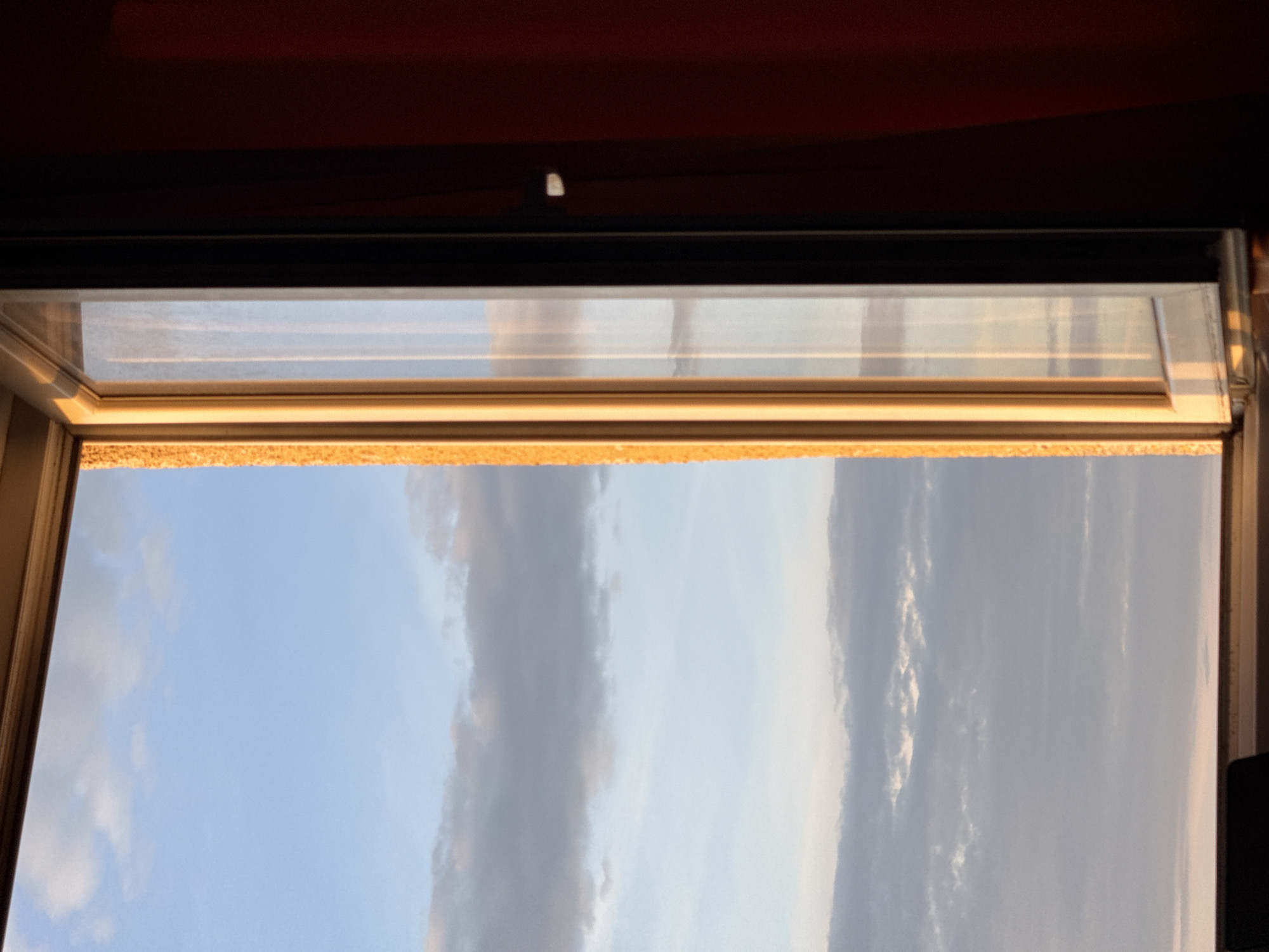 The image shows a view of the sky through an open window or skylight. The sky is blue with various cloud formations, some appearing elongated and vertical. The window frame is visible at the top and sides, appearing to be made of dark metal and wood. The interior of the room is visible at the very top of the frame, appearing dark. Based on the architectural style and the possible presence of Velux windows in the scene, it may be inside a building within the city of Sofia, Bulgaria.