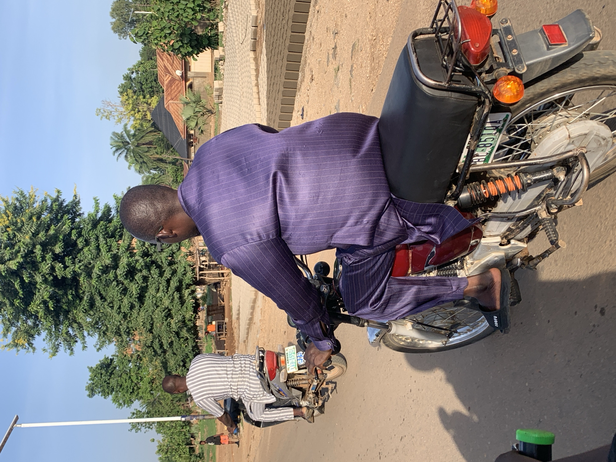 A man in a purple striped suit rides a motorcycle on an asphalt road. The motorcycle has a license plate that reads "HC-85174 LA-10-5501." Another motorcycle, partially visible in the lower portion of the image, has a rider in a striped shirt. They are riding on an asphalt road. Buildings and greenery are visible in the background. Trees are on the left and the edge of a brick walkway, and a building is on the right. The photo was taken in the city of Maigizo, Nigeria.