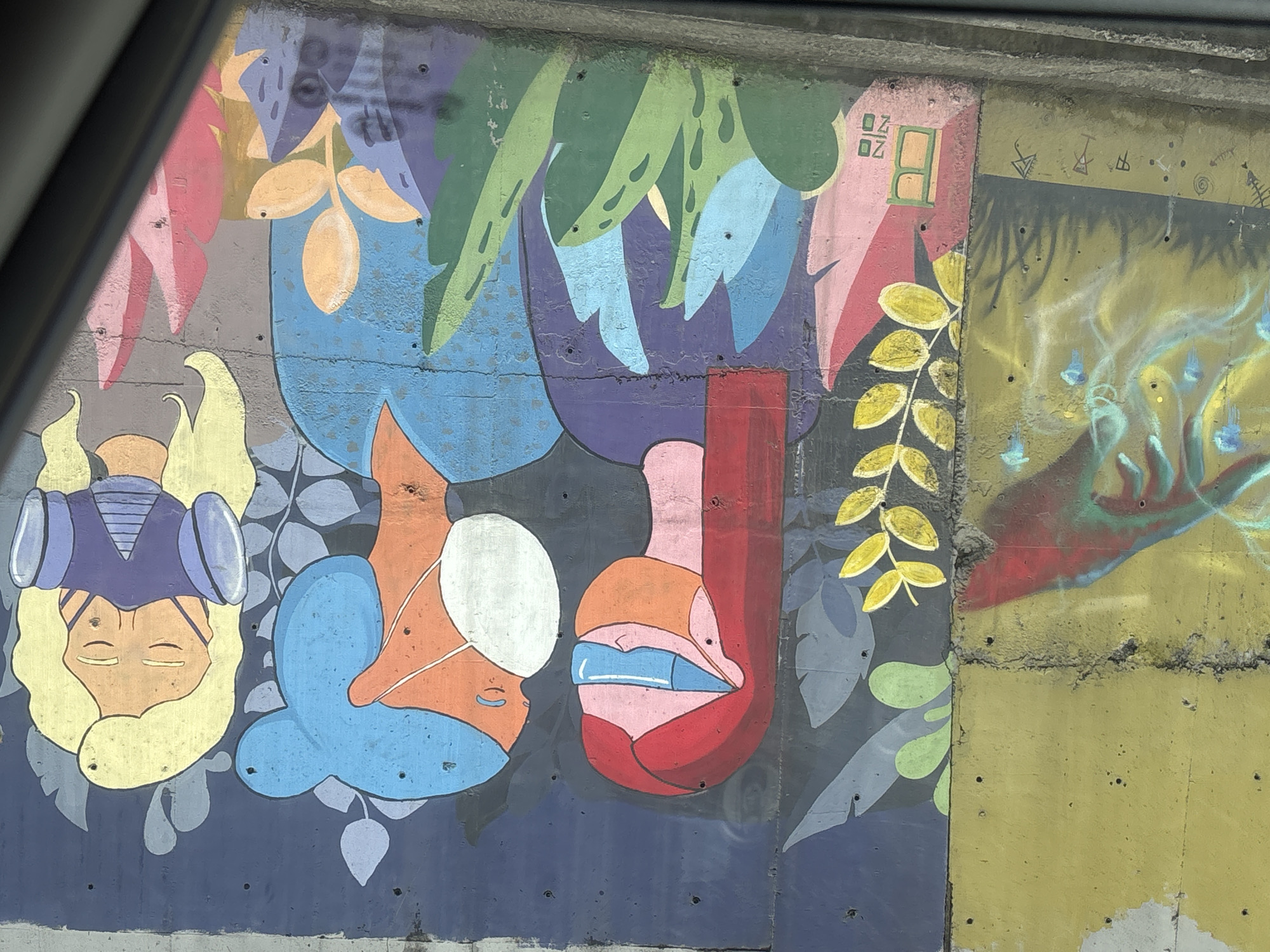 An outdoor concrete wall in Tbilisi, Georgia, displays two distinct sections of graffiti art. The larger right section features a vibrant mural on a dark blue background depicting three stylized human-like figures. From left to right within the mural, the first figure has red hair, wears a purple top, an orange face mask, and blue goggles. The central figure has short blue hair/hat, wears a blue top, and a white medical-style face mask; this figure faces the red-haired individual. The third figure, on the right, has blonde hair, wears a light purple top, and a dark purple respirator mask. All three figures have closed eyes. The lower portion of the mural is adorned with various stylized plant motifs in shades of green, blue, pink, and orange. A signature "BOZO" is visible vertically in green letters on a pink and orange element at the bottom-left of the mural section. The concrete wall displays minor imperfections and small drill holes. To the left, separated by a vertical seam in the concrete, a smaller section of the wall is painted yellow and features abstract graffiti, including a red and turquoise-green design with glowing accents and small symbols below it. A dark, diagonal object, likely a vehicle frame, partially obstructs the view from the bottom-right corner.