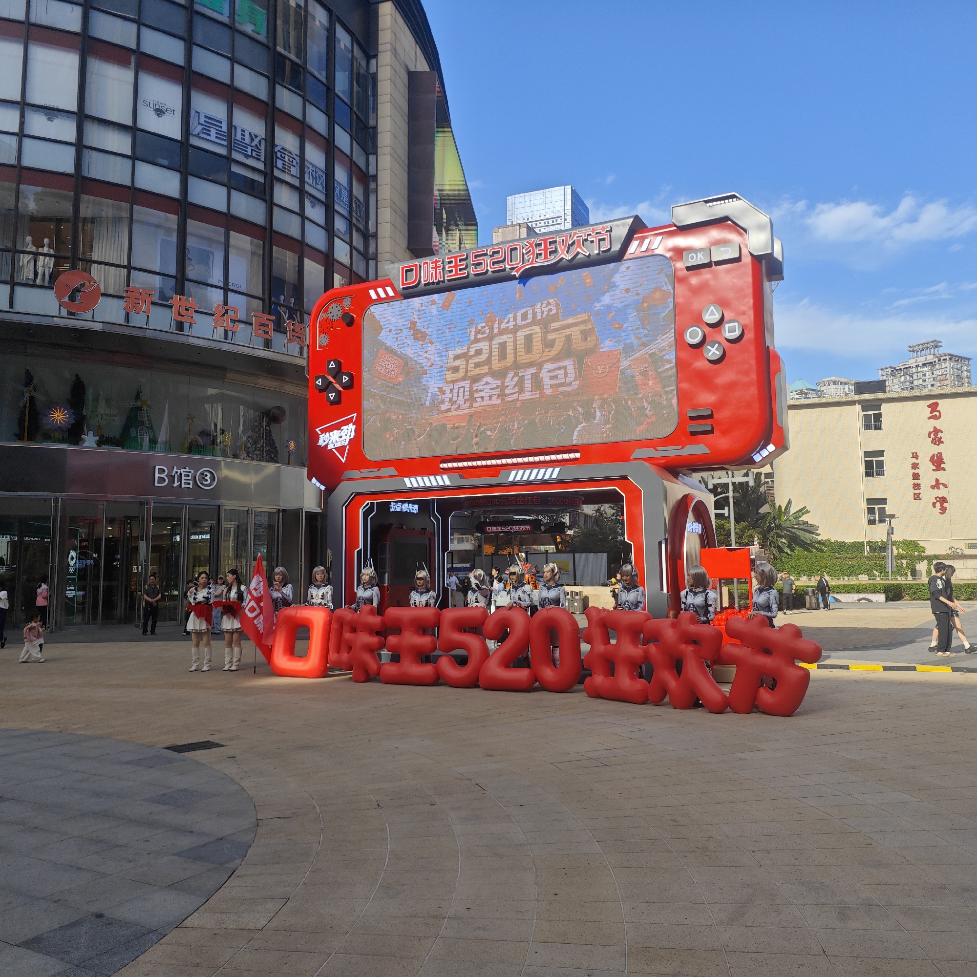 The image shows a promotional event held in a public space. A large, red gaming console-shaped structure serves as a stage and display. It features a screen showing promotional content and is adorned with gaming controller buttons. In front of the structure are inflated, large red Chinese characters. A group of individuals dressed in matching costumes, possibly for promotional purposes, stand near the structure. Other people are also seen walking nearby. In the background, a multi-story building with glass windows and commercial signage is visible, along with another building that has "马家堡小学" in red letters, which translates to "Majiapu Primary School," indicating the Yuzhong District, China. The event is taking place on a paved public square with a patterned ground. The sky is blue.