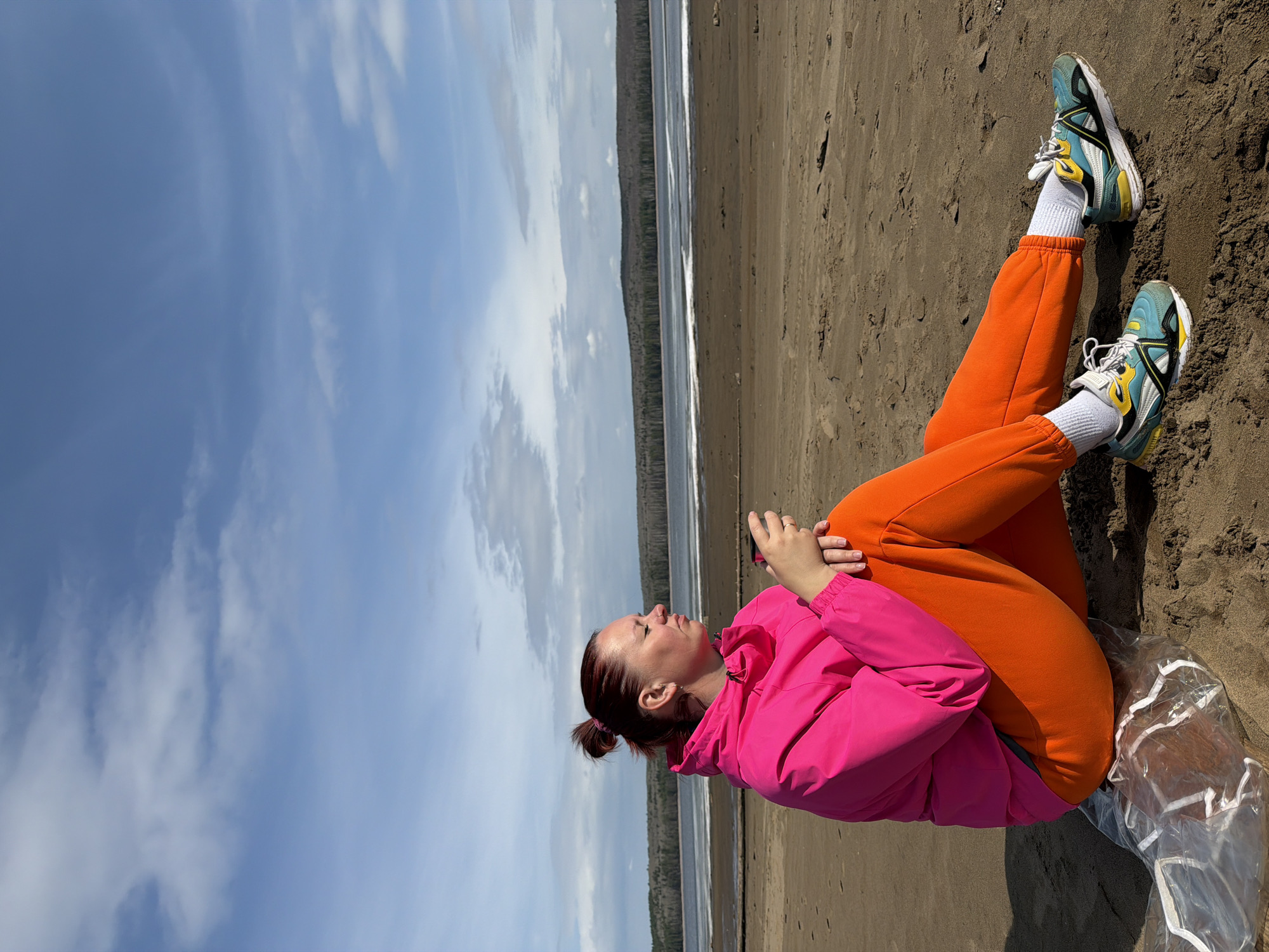 The image depicts a woman sitting on a sandy beach on what appears to be a sunny day. The woman has reddish-brown hair pulled back in a bun, and she is wearing a bright pink jacket and orange sweatpants. She also has on white socks and light blue sneakers with yellow accents. The woman seems to be looking up and slightly to her left, with her hands clasped in front of her. The sand beneath her appears to be coarse and brown. Behind her, a line of trees is visible, followed by a body of water. The sky is blue with scattered white clouds.
While the prompt mentions Chaikovsky, Russia, without additional information, I cannot pinpoint the exact location on the shore.