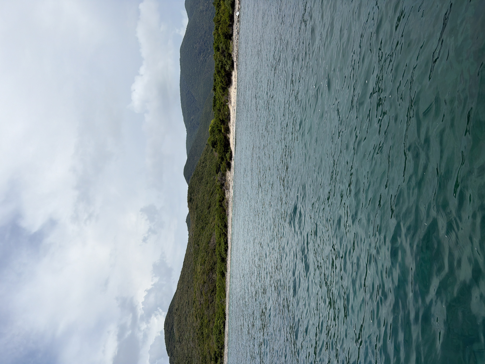 The image shows a coastline with lush, green mountains meeting a body of water, likely the sea. The water's surface is rippled. A narrow beach is visible between the mountains and the water. The sky is overcast with scattered clouds. The location appears to be near a tropical or subtropical area, possibly in Nha Trang Ward, Vietnam, judging from the terrain and vegetation. There are no people visible in the image.
