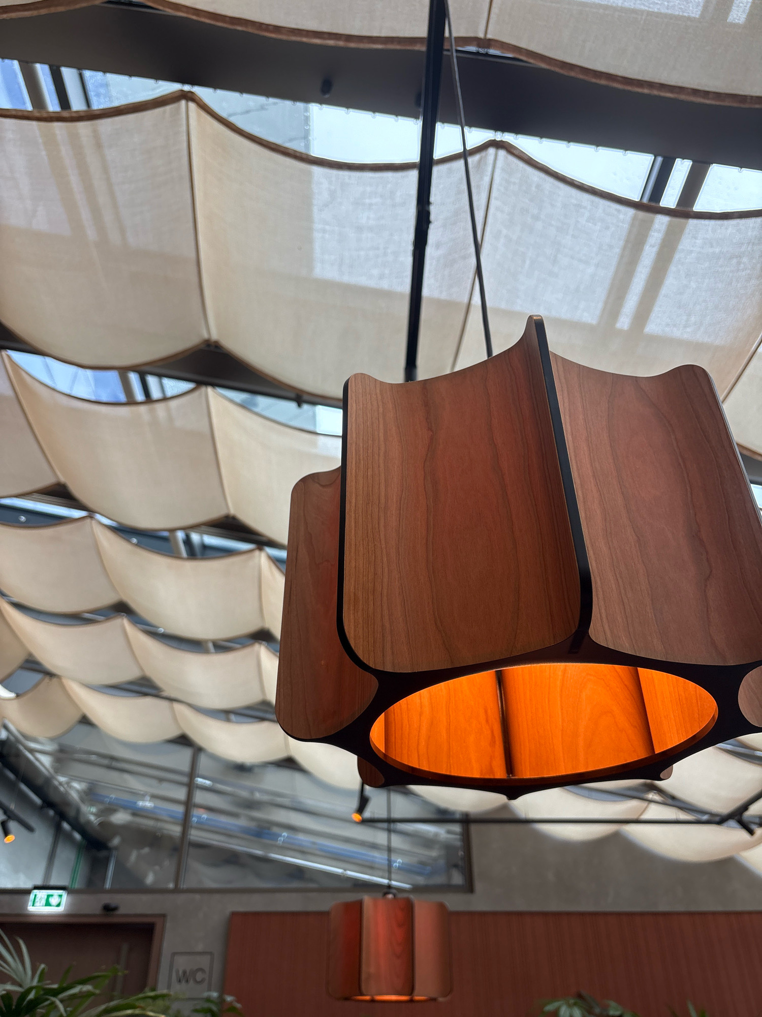 The image shows an interior view of a building, likely a restaurant or cafe, with a distinctive wavy ceiling made of beige fabric panels stretched over a metal frame. A prominent wooden pendant light fixture with a warm orange glow hangs in the foreground. In the background, through large windows, there is a glimpse of a city environment with buildings and possibly some industrial elements. A "WC" sign is visible on a wall, indicating a restroom facility. The overall impression is one of modern, stylish interior design. The location in Kopavogur, Iceland, suggests a contemporary establishment in a Scandinavian setting. There are no people or animals visible in the image.