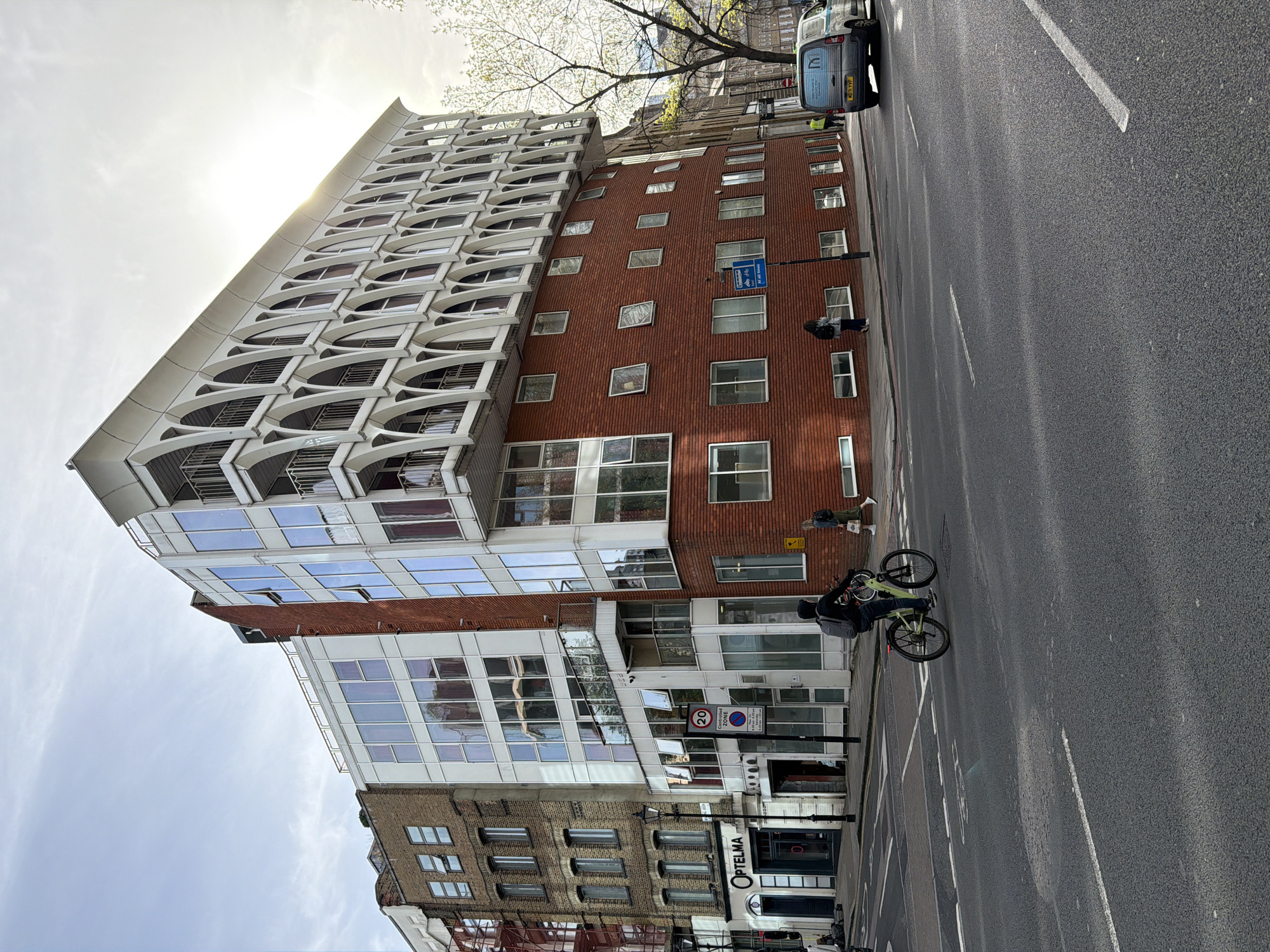 The photograph captures a street scene in London, United Kingdom. The dominant architectural feature is a tall building comprised of distinct sections: a lower brick portion, a mid-section of glass and brick, and an upper section with a white facade featuring curved design elements over the windows. The lower part of the building is adjacent to an older brick building with smaller windows.
The street is asphalt with painted white lane markers. A cyclist is in the foreground and there are a pedestrian and a car in the background. A few street signs are visible near the base of the building. The lighting suggests it is daytime, with the sun partially obscured by the buildings. The sky is visible in the upper portion of the image.