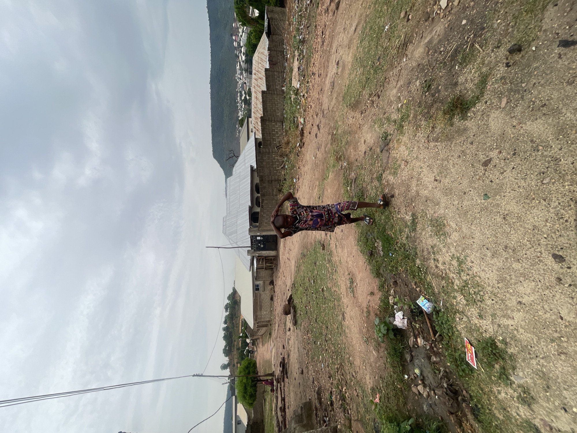 A person stands on a dirt path near houses in what appears to be a settlement in Lokoja, Nigeria. The person is wearing a patterned shirt and shorts and has their hands behind their head. The dirt path has some grass and trash on it. The houses are simple structures, and a hillside is visible in the distance. The sky is overcast.