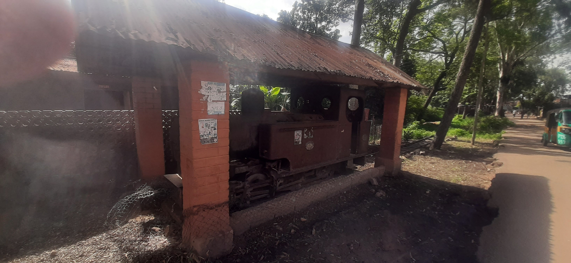 The image captures a view of an old, rusty locomotive engine housed under a weathered tin roof structure, supported by brick pillars. The structure appears to be situated in an outdoor area, possibly a historical site or a disused railway station in Jos, Nigeria. Vegetation, including trees and foliage, surrounds the area, with a dirt road visible to the right, where a green tuk-tuk vehicle is parked. The scene is illuminated by sunlight, casting shadows and creating a somewhat hazy atmosphere. There are posters attached to one of the brick pillars. The overall impression is one of a forgotten piece of history in a lush, semi-tropical environment.