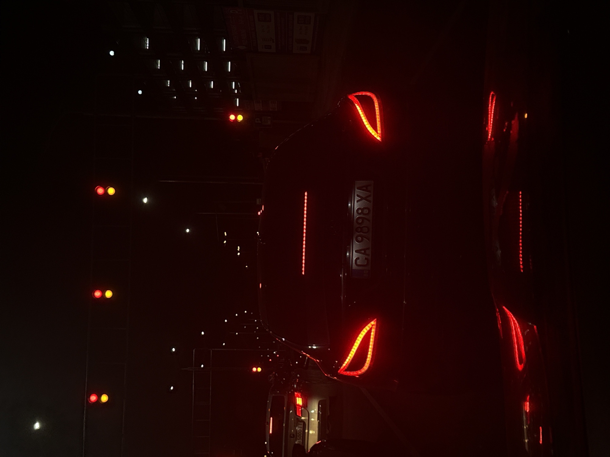 The image depicts the rear of a car with Bulgarian license plate "CA 9898 XA" at night in Plovdiv, Bulgaria. The car's taillights are illuminated, as are various streetlights in the background. The overall lighting is dim and dominated by red and orange hues. Vertical structures, possibly streetlights or buildings, are visible in the background. The orientation of the image is rotated, as the top of the car is oriented to the left side of the frame.