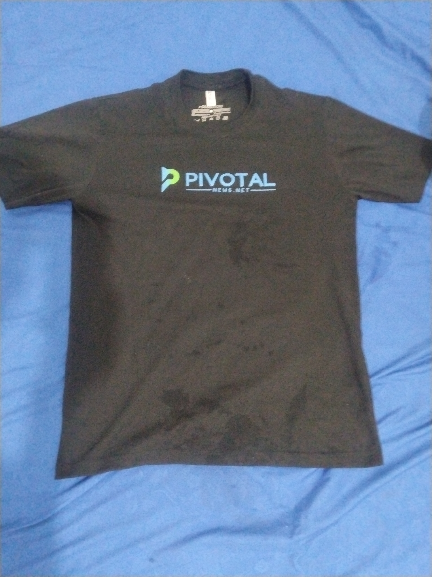 A black t-shirt with the "Pivotal News Net" logo in light blue and green is lying on a blue fabric surface. The shirt appears slightly wrinkled and may have some stains or faded areas. The setting appears to be a personal or domestic one given the unadorned backdrop, but no specific location within Lhokseumawe, Indonesia can be determined from the image. There are no people or interactions visible in the image.