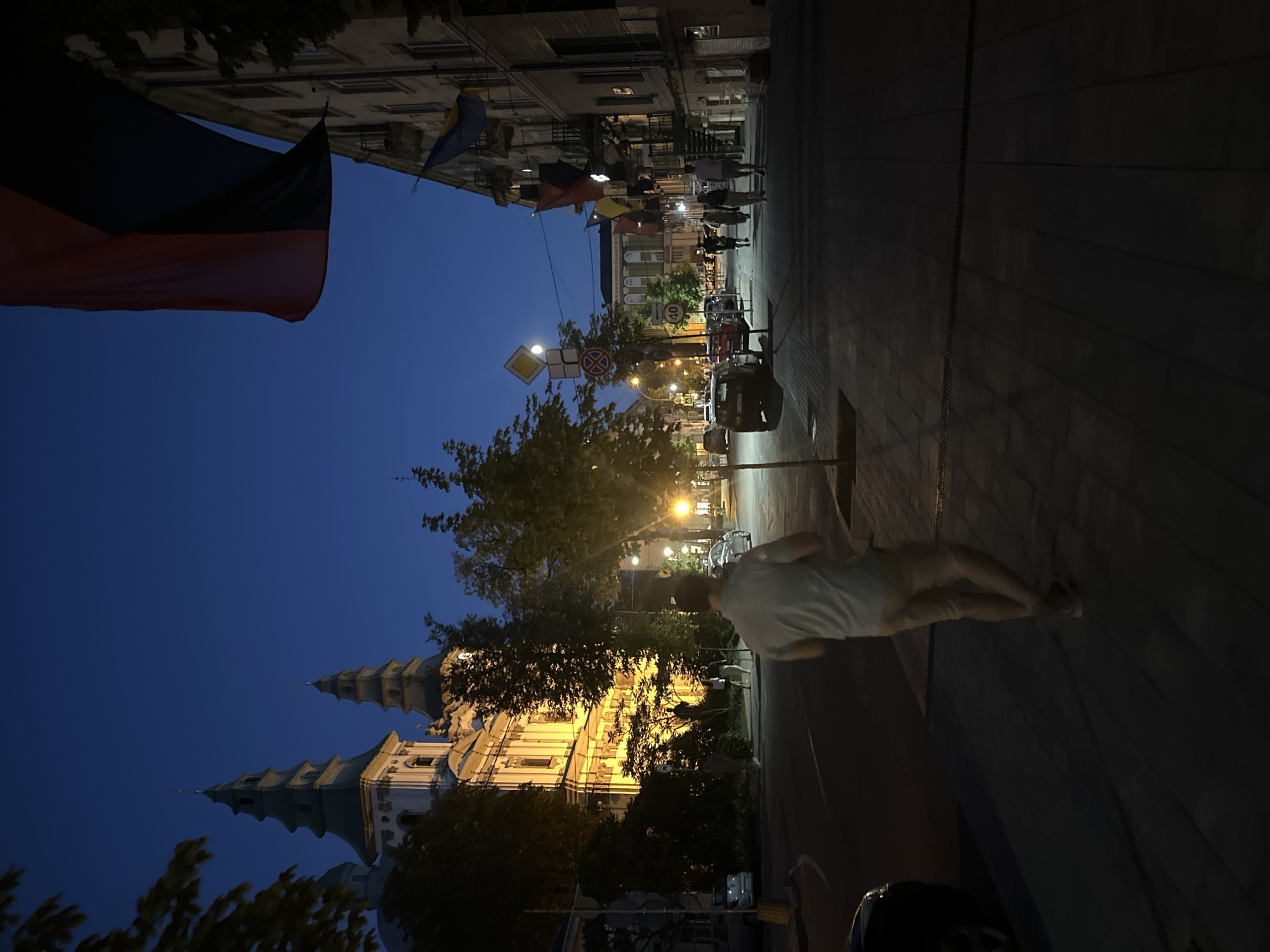 The image depicts a city street scene in Ternopil, Ukraine, at dusk or early night. The sky is a deep blue, transitioning from the fading daylight. On the left side of the street, a grand, brightly lit building with multiple spires, resembling a church or a historical architectural landmark, is illuminated by warm yellow lights, casting a glow onto the surrounding area. Trees with dark foliage are positioned in front of and around the building.
The street itself is paved with dark rectangular stones. On the right side of the street, a multi-story building with balconies lines the sidewalk. Several flags, in various colors including red, black, blue, and yellow, are hanging from the building or along the street. Some are large, draped flags, while others appear to be smaller banners.
Several people are visible walking on the sidewalk. In the foreground, a person wearing a light-colored t-shirt and beige shorts is walking away from the camera, towards the right side of the image. Further down the street, more figures are walking, some in groups.
Cars are parked along the street and driving on the road. Traffic signs, including a speed limit sign showing "40" and a diamond-shaped yellow sign, are visible. Streetlights are on, casting pools of light on the pavement and illuminating parts of the scene. The overall atmosphere is calm and urban, with the illuminated architecture and flags suggesting a sense of place and possibly civic pride.