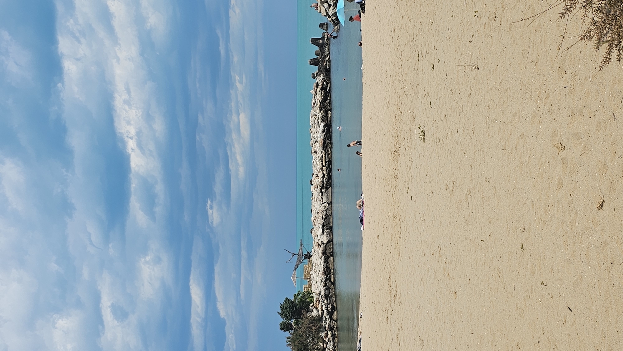 This image shows a beach scene in Varna, Bulgaria. The scene is split horizontally between the sky/sea on the left and the sand on the right. A rocky breakwater extends into the sea, defining a lagoon-like area with people swimming and standing in the water. On the beach, the sand is light-colored, with sparse vegetation. In the distance, vegetation and structures are visible along the breakwater. The sky is blue with layered clouds.