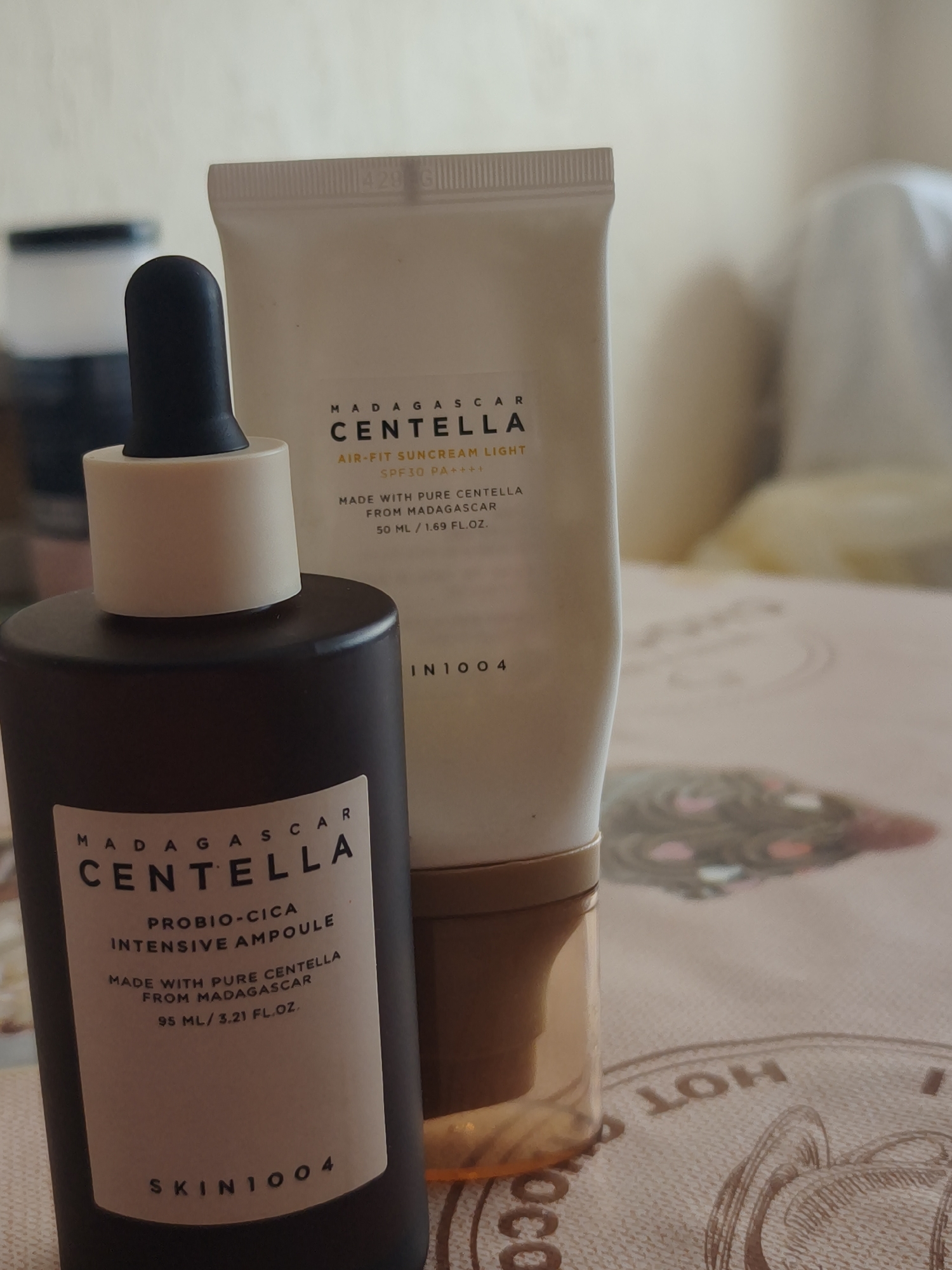 Two skincare products are displayed on a patterned tablecloth. The product in the foreground is a dark brown bottle with a dropper dispenser, featuring a label with white text. The label reads "MADAGASCAR CENTELLA PROBIO-CICA INTENSIVE AMPOULE MADE WITH PURE CENTELLA FROM MADAGASCAR 95 ML / 3.21 FL.OZ. SKIN1004". Behind this bottle is a white tube with a gold base, also featuring text on its label. This text includes "MADAGASCAR CENTELLA AIR-FIT SUNCREAM LIGHT SPF30 PA++++ MADE WITH PURE CENTELLA FROM MADAGASCAR 50 ML / 1.69 FL.OZ. SKIN1004". The background is blurred, suggesting an indoor environment. The lighting is soft and diffused.