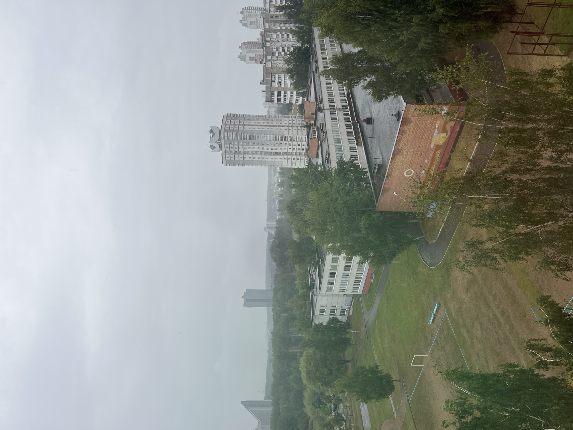 The image depicts an overcast day in Minsk, Belarus, with a view of an urban landscape dominated by residential buildings, trees, and a sports field. In the foreground, a sports field with a soccer goal and two blue benches is visible, surrounded by a lawn and trees. Behind the field, a low-rise brick building with a mural on its side is situated. Further in the background, several multi-story apartment buildings, including a distinctive tall white building with a rounded top, are scattered across the horizon. A large expanse of green trees separates the closer buildings from the distant ones. The sky is a uniform gray, suggesting cloudy or rainy weather. The overall impression is one of a residential district on a subdued, possibly damp day.