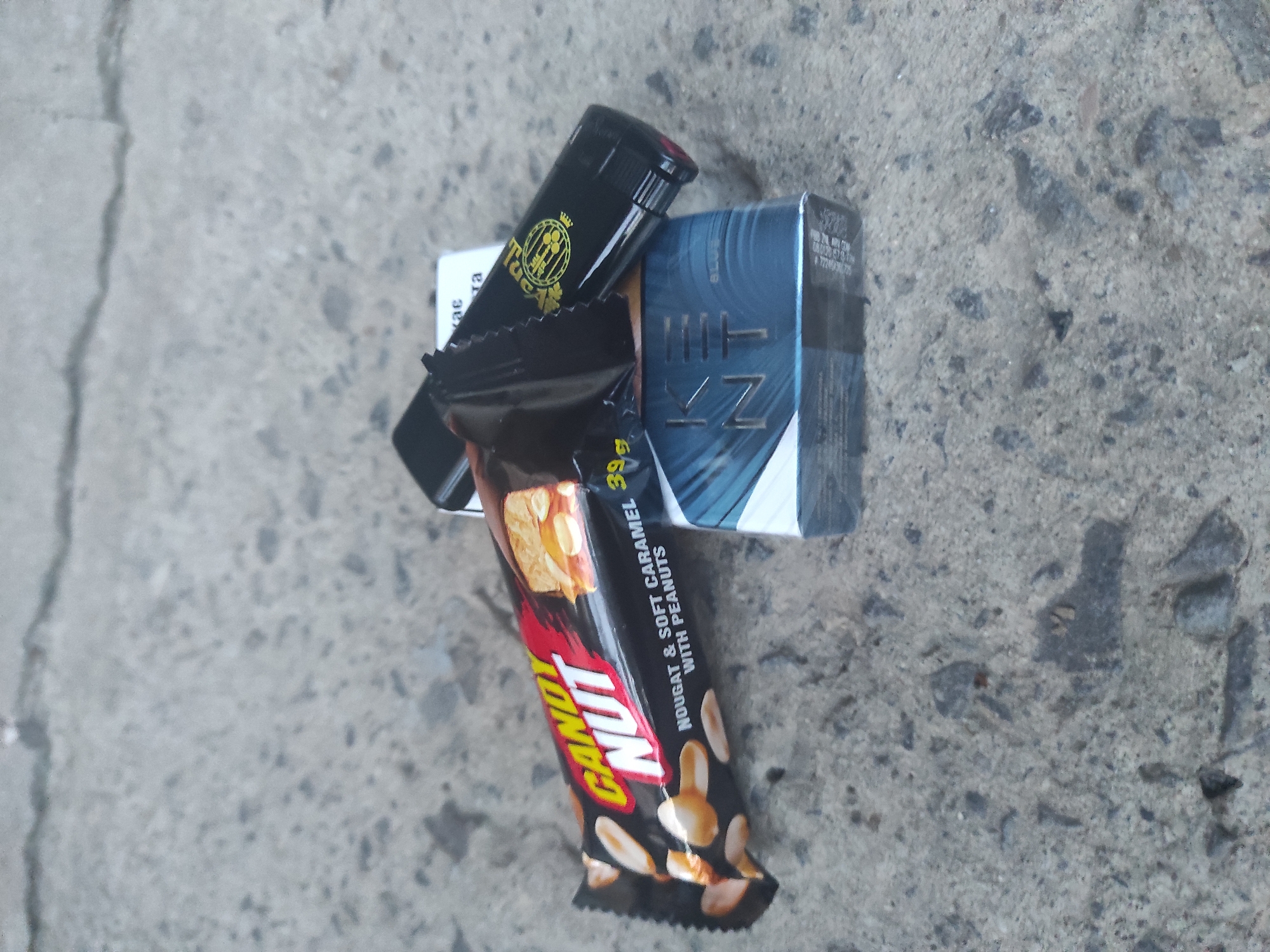 The image displays a close-up overhead view of several items resting on a light gray concrete surface, possibly a sidewalk or pavement. The texture of the concrete is rough, with small stones and cracks visible.
The main subjects are a black lighter, a blue-green cigarette pack, and a black and orange candy bar wrapper. The items are arranged somewhat haphazardly; the lighter is on top of the cigarette pack, which is partially obscured by the candy bar lying across it.
Visible text on the items includes:
- On the black lighter, a gold logo with the word "Tucson" and a crown/crest design above it.
- On the cigarette pack, the brand name "KENT" is clearly legible in white capital letters against the blue-green background. Smaller, illegible text is present on the side of the pack.
- On the candy bar wrapper, the brand name "CANOT NUT" is prominent in yellow and red lettering. Below it, the description "NOUGAT & SOFT CARAMEL WITH PEANUTS" is visible. The weight "39 g" is also present on the wrapper.
There are no people or explicit activities depicted. The scene appears to be outdoors in daylight, suggested by the clear and even lighting, but no specific time of day or weather conditions can be determined. The concrete surface provides the only location cue, indicating a generic outdoor setting. No particular emotions are conveyed by the image.