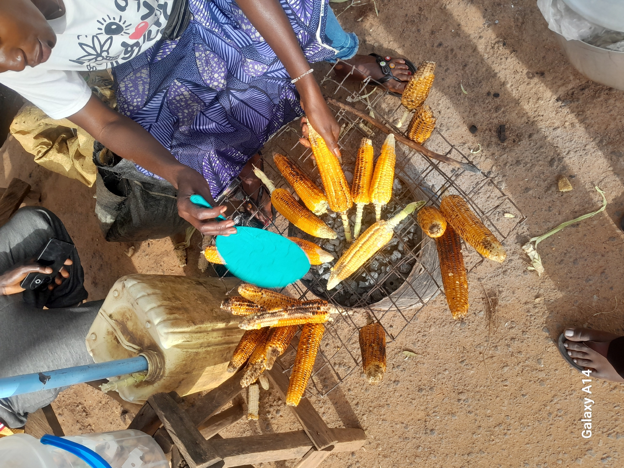 The image depicts an outdoor scene, likely in the city of Barakin, Nigeria, where roasted corn is being prepared and sold. A woman, wearing a white t-shirt with a black graphic and a patterned purple and blue skirt, is actively engaged in the process, holding a blue tool over a grill laden with corn on the cob. Several cobs of corn, some partially roasted and others more fully cooked with visible char marks, are arranged on a metal grill over what appears to be an open fire or coals. The ground is sandy and dusty, typical of outdoor market environments. In the background, a large, yellow plastic container with a blue spout stands on a wooden crate. Another person, whose legs and lap are visible, is seated nearby, holding a black smartphone. The overall atmosphere suggests a bustling street food vending operation. The presence of the "Galaxy A14" watermark indicates the device used to capture the image.