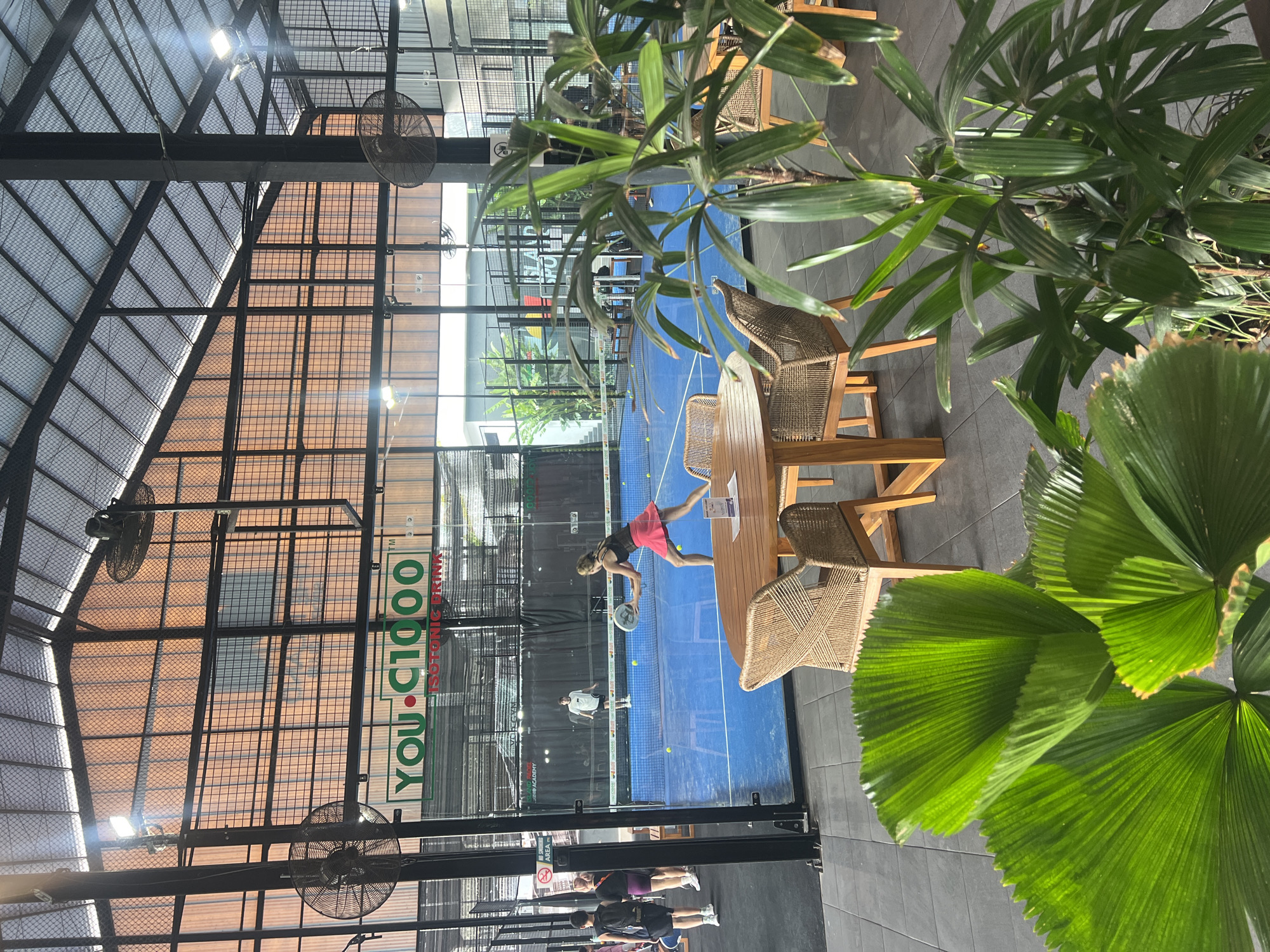 The image depicts an indoor padel court in North Kuta, Indonesia. The court has a blue surface and is enclosed by glass walls. Two individuals are actively playing padel on the court. One player, a woman in a pink skirt, is in the process of hitting the ball. Another player, a man in a white shirt and shorts, is positioned on the opposite side of the net. In the foreground, a wooden table with woven chairs is set up, suggesting a seating area for spectators or patrons. Lush green plants are positioned to the right of the frame, adding a natural element to the indoor environment. The background reveals the architectural features of the facility, including a high ceiling with exposed beams and industrial-style fans. A prominent banner with the "YOU C1000" logo is visible above the court. The overall scene captures a recreational sporting activity within a modern indoor complex.