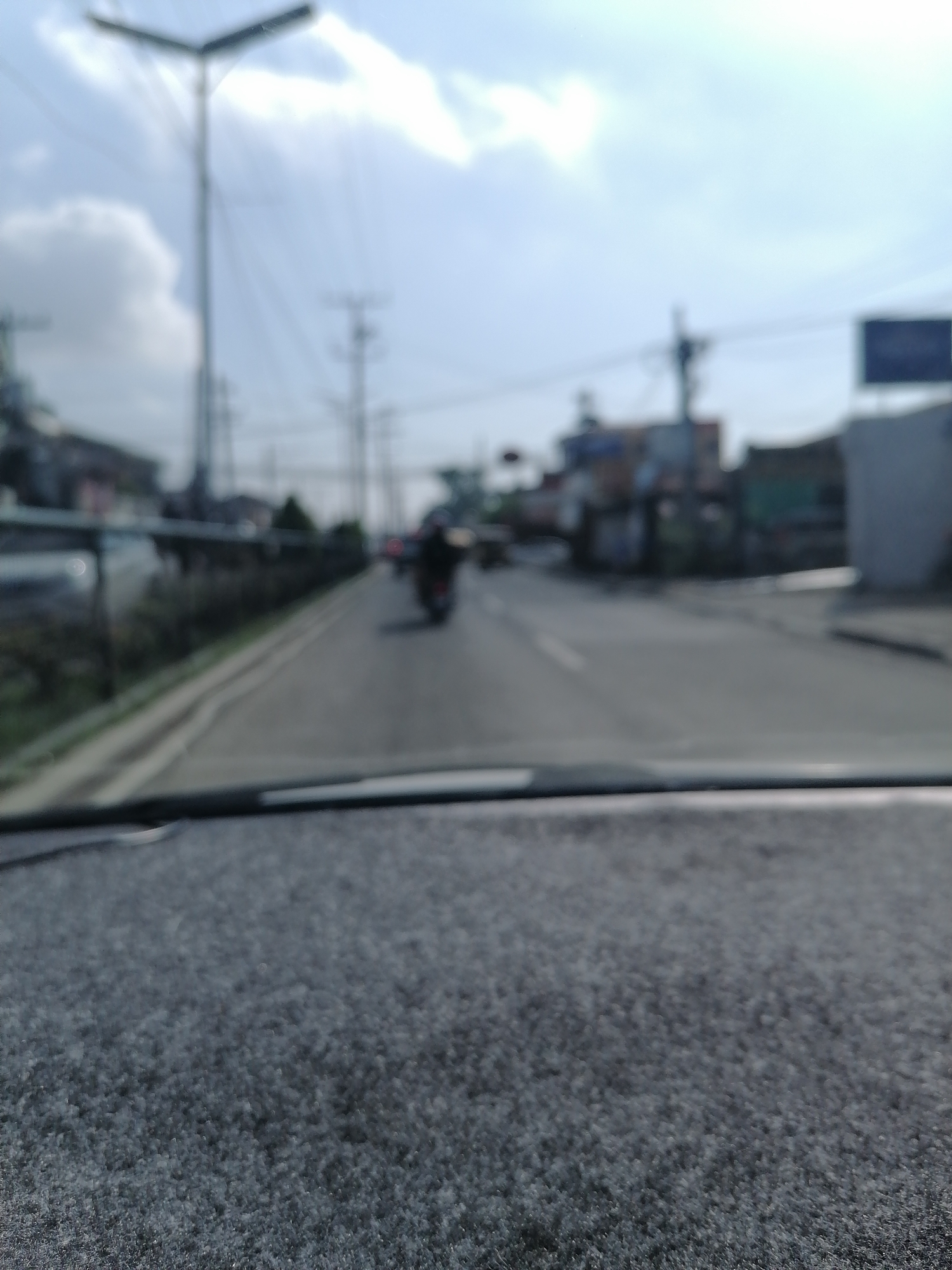 The image captures a heavily blurred, daytime street scene from the perspective of inside a vehicle, looking out through the windshield. The foreground is dominated by the textured, dark grey surface of what appears to be a car dashboard or carpet.
Beyond the dashboard, a road or street stretches into the distance, appearing wet or reflective in areas. A faint white line is visible marking the center or edge of the road. On the left side of the road, there's a sidewalk or shoulder bordered by a low barrier and some indistinct vegetation. In the mid-ground, a dark shape resembling a person on a motorcycle is vaguely visible, moving away from the viewer.
The background consists of a mix of urban and natural elements under a bright sky with scattered white clouds. Several utility poles with power lines crisscross the frame. A prominent street light with a V-shaped fixture stands tall on the left. On both sides of the street, blurred outlines of buildings, some with multiple stories, are discernible. On the far right, a dark, rectangular sign is visible against the sky, but no text is legible due to the extreme blur. The sun appears to be somewhat high in the sky, creating a bright wash, suggesting a sunny day.
The overall impression is one of movement or a missed focus, providing a soft, impressionistic view of a bustling street in what is indicated to be Davao City, Philippines. No specific text or distinct emotional cues are present.