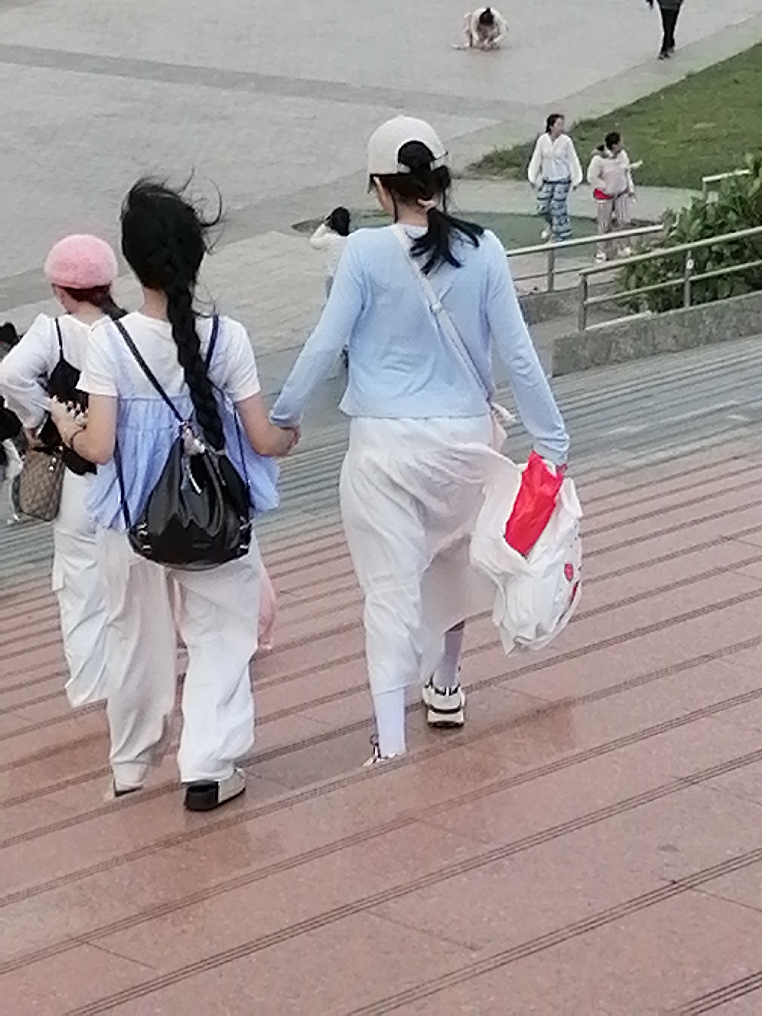 The image depicts a group of people on a sloped, paved surface that appears to be outdoor stairs. In the foreground, three young women are walking up the stairs. The woman on the left wears a white top with a black strap, white pants, and a pink beret. She is carrying a patterned shoulder bag. The middle woman has long black hair in a braid, wears a light blue top over a white top, and white pants. She is carrying a large black bag on her back. The woman on the right wears a light blue long-sleeved shirt and white pants. She is holding a white plastic bag with red items inside. In the background, several other individuals are visible, some walking and one person kneeling. The environment appears to be a public outdoor space, possibly a park or promenade, with grassy areas and metal railings visible. There is no information in the image to suggest the location is Da Lat, Vietnam.