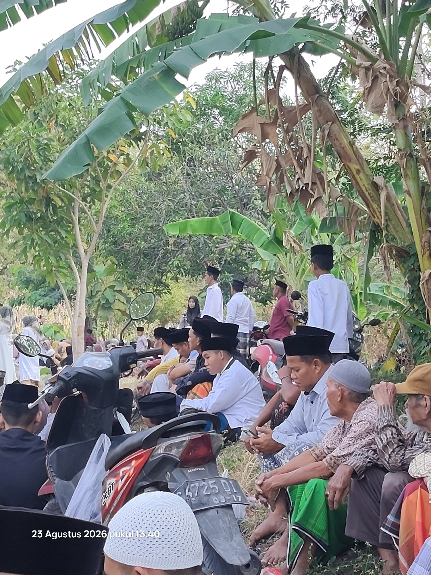 In a rural outdoor setting, likely in Padang Mentoyo, Indonesia, a group of men are gathered, some seated on the ground and others on or near motorcycles. The scene is characterized by lush greenery, with prominent banana trees and other foliage. The men are dressed in traditional Islamic attire, predominantly white shirts and black caps (peci).
In the foreground, a motorcycle with a black seat and red accents is parked. Another motorcycle, also black, is partially visible. Several men are seated in front of these motorcycles, facing to the right. One man in the immediate foreground wears a black cap and a dark garment. To his right, a man in a light blue shirt and sarong is holding a smartphone. Further to the right, an older man with a patterned shirt and a grey cap is visible, and next to him, another man wears a tan cap and an orange garment.
In the middle ground, more men in white shirts and black caps are seated or standing. One man in a white shirt stands near a motorcycle, looking to the right. A woman in a hijab is partially visible in the background. On the left side of the image, a few individuals, including women, are dressed in white attire and appear to be standing.
The environment suggests a warm climate, with the sun casting shadows. The overall impression is that of a community gathering, possibly for a religious or cultural event. The date stamp in the bottom left corner reads "23 Agustus 2026 pukul 13.40".