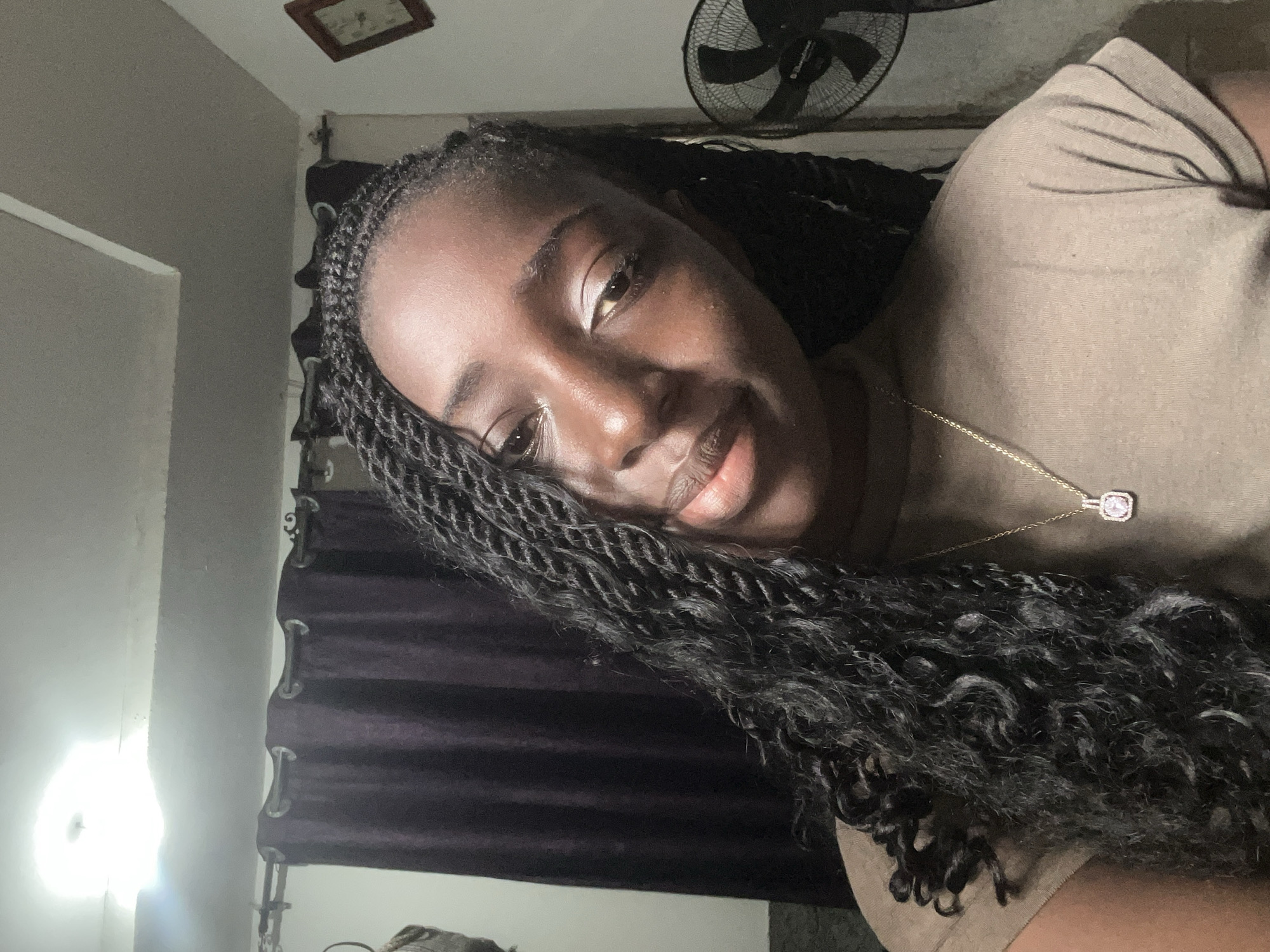 The image is a medium shot of a young Black woman with braided hair and a light pink pendant necklace. She is looking slightly away from the camera with a relaxed expression. She is wearing a taupe t-shirt. The background appears to be an indoor setting, possibly a bedroom or living room, with dark purple curtains, a ceiling light, and a black fan visible on the right side. There is also a framed picture on the wall. The lighting suggests it may be evening or indoors with artificial light. The scene is a casual, personal moment captured in what seems to be an urban environment, potentially in Abuja, Nigeria, given the context provided.
