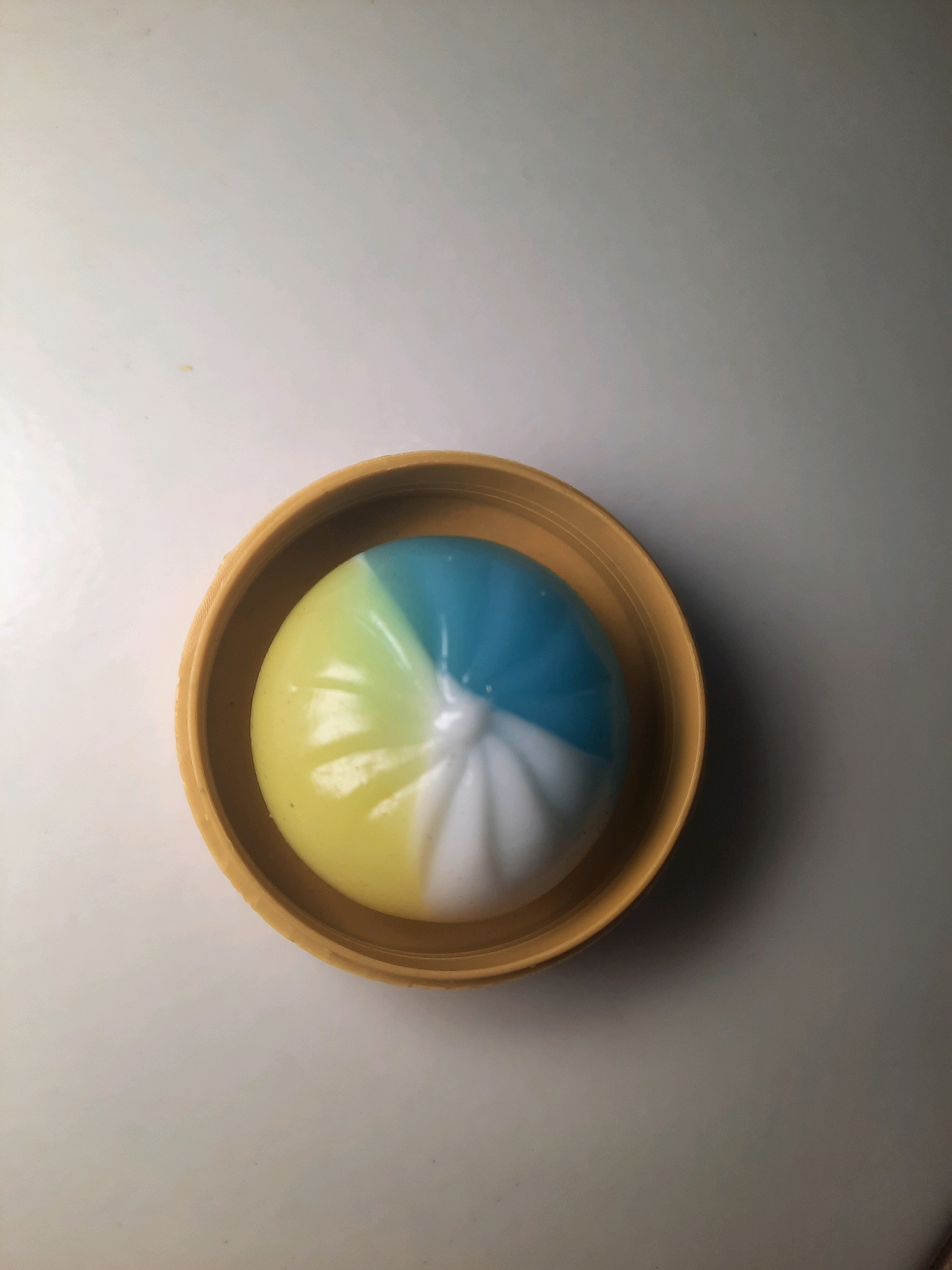 A colorful, spherical stress ball, divided into yellow, blue, and white sections, rests inside a golden-colored shallow bowl. The bowl is placed on a white, flat surface.