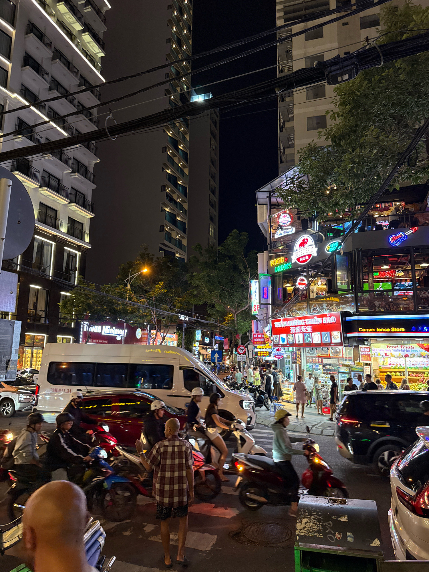 The image depicts a bustling street scene at night in Nha Trang, Vietnam. The foreground is dominated by a busy intersection with several motorcycles and cars. A man in a red and white plaid shirt stands facing away from the camera, gesturing with his right hand, as if directing traffic or interacting with someone off-camera. Several other individuals are seen riding motorcycles, some wearing helmets. A white van is parked in the middle of the road.
The background features tall, modern buildings illuminated by the night. One building on the left has a white facade with balconies and strip lighting along its edges. Another building to its right is taller and appears to be under construction or has a darker, more industrial aesthetic. Along the street level, a vibrant commercial area is visible, with numerous signs and storefronts displaying bright, colorful lights. These include businesses labeled "FOOD & DRINKS HUB," "Convenience Store," and what appears to be a fruit stand or market selling fresh produce. The overall atmosphere is one of urban nightlife and activity.