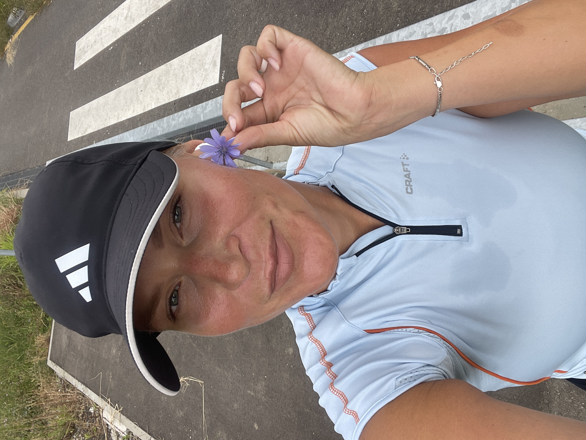 A woman wearing a black Adidas baseball cap and a light blue athletic shirt holds a small purple flower up to her cheek. Her shirt appears damp, suggesting she has been exercising. She is standing on a paved surface with white stripes, resembling a crosswalk. The background shows a grassy area and a road. The scene is outdoors. No specific location in Sochi, Russia, can be identified from the image.