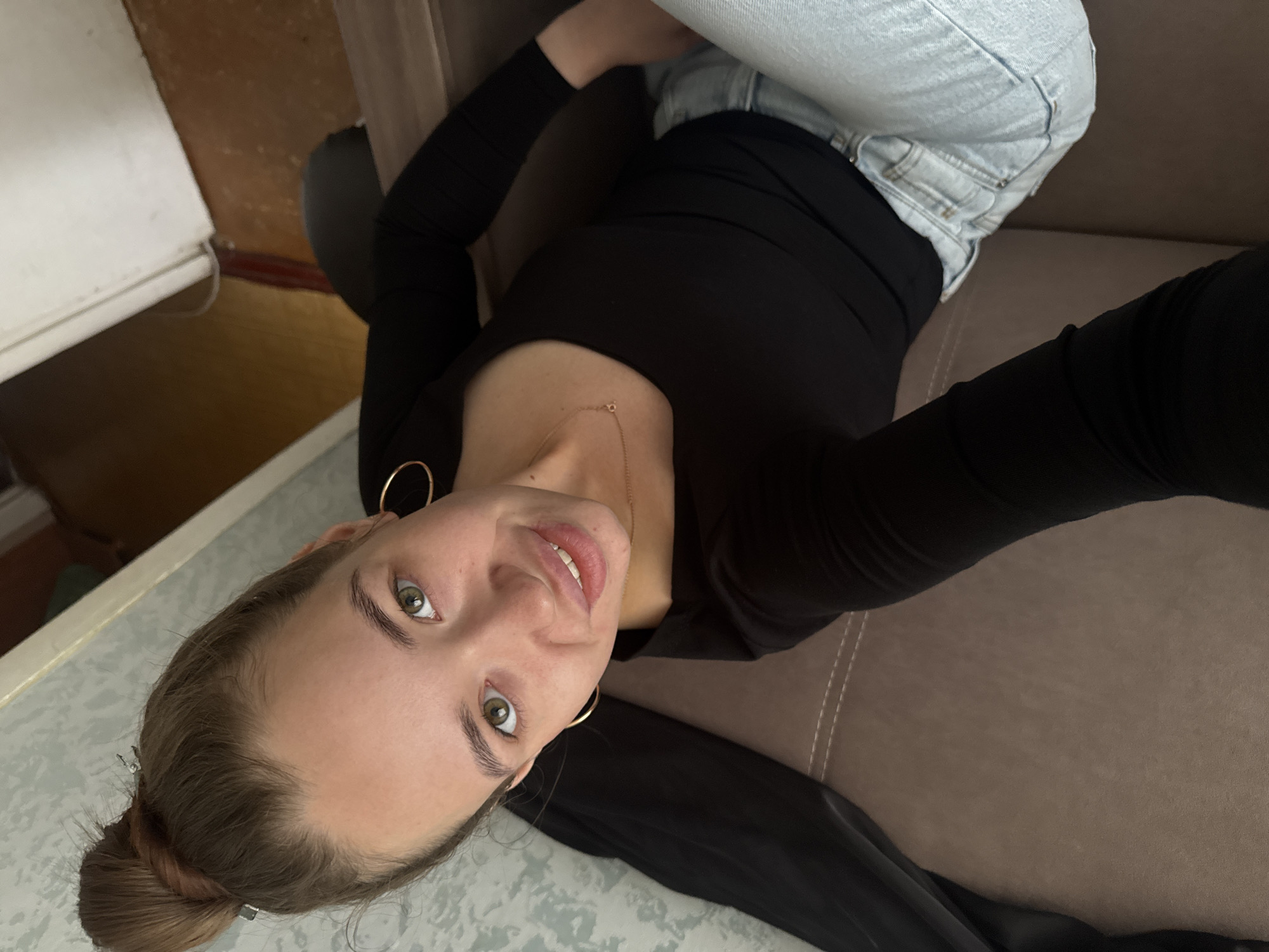 In a dimly lit indoor setting, a young woman with her hair styled in a bun is captured from a high-angle perspective, taking a selfie. She is seated on a brown sofa, wearing a black long-sleeved shirt and light blue jeans. The woman has green eyes and is smiling at the camera. A delicate gold necklace adorns her neck, and she wears hoop earrings. The background consists of a textured wallpaper on the left and a floor and door frame on the right, suggesting an interior of a residential dwelling. The image does not provide specific visual cues to pinpoint the exact location within the city of Chaikovsky, Russia.