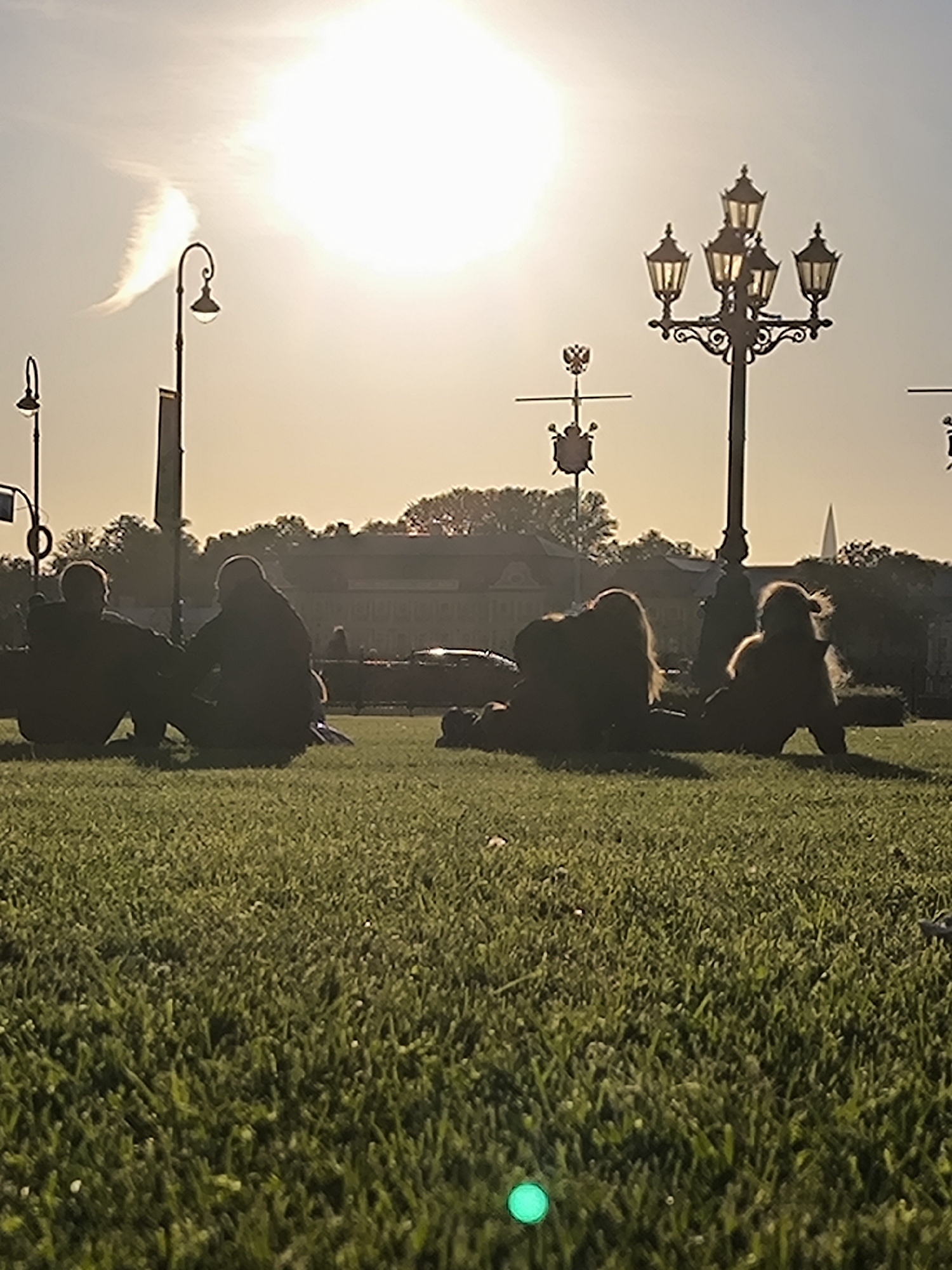 In a park setting with a grassy lawn, four individuals are seen from the waist down, positioned on the grass. Two figures are seated next to each other in the mid-ground on the left, facing away from the camera. To the right of them, also in the mid-ground, two other figures are lying down or reclining on the grass. The foreground of the image is dominated by lush green grass, with a bright green circular object visible in the lower center. The background features a clear sky with a bright sun and some wispy clouds. A streetlamp with multiple lights is prominently positioned in the upper right quadrant of the image. In the far background, a row of buildings and trees can be observed. The overall scene is captured during daylight with the sun casting a bright glow.