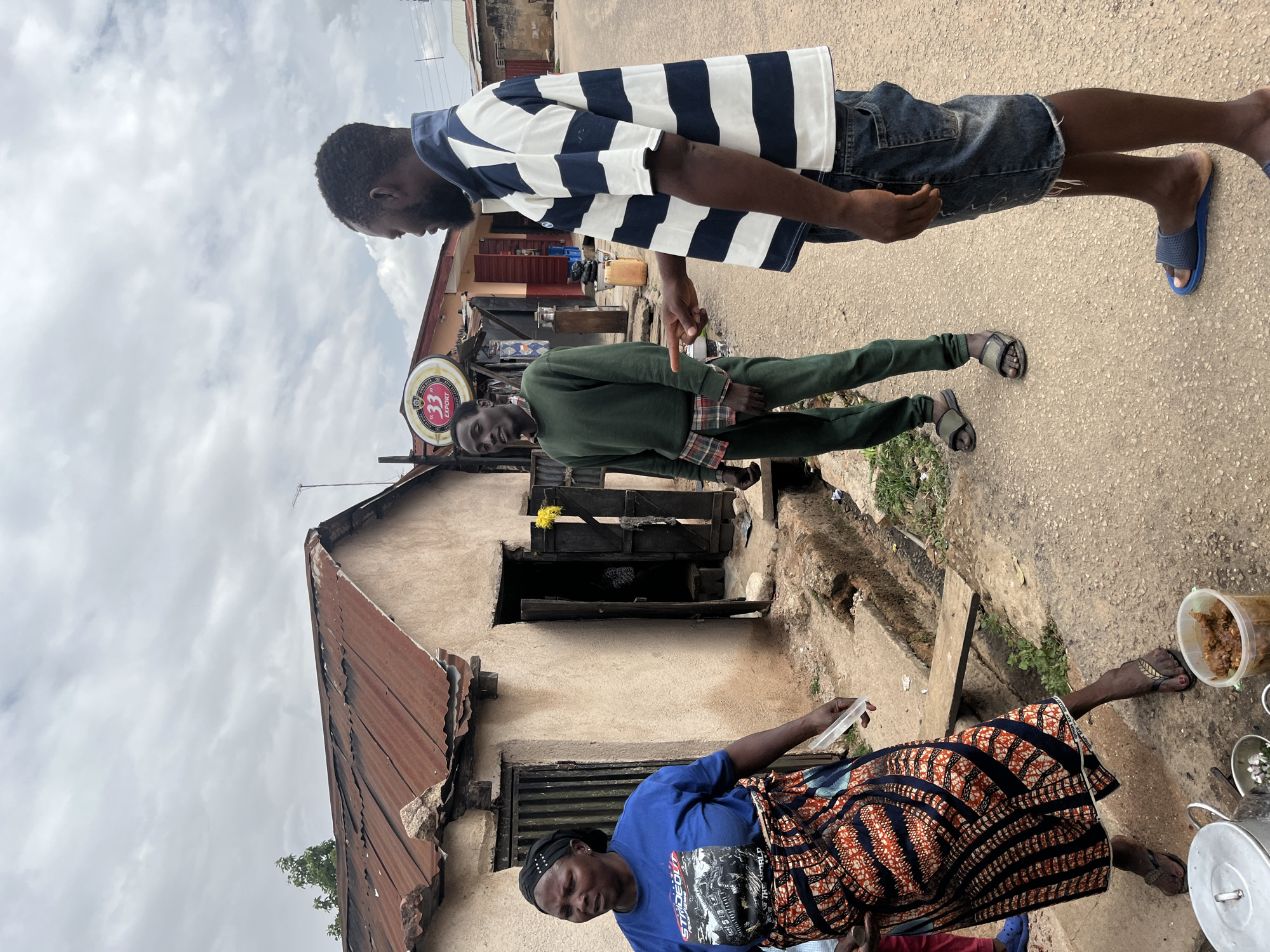 The image, taken in Jos, Nigeria, depicts a street scene with three individuals and a building. A woman wearing a blue shirt and an orange and black patterned skirt sits near cooking pots. She appears to be engaged in preparing or selling food. Behind her is a weathered building with a rusty corrugated metal roof. Two men stand in the street. One wears a striped shirt and denim shorts, while the other is dressed in a green outfit. The man in the striped shirt is gesturing towards the man in green with his hand. A beer sign is visible attached to the building, but it’s not legible. The street is unpaved. The weather appears overcast.