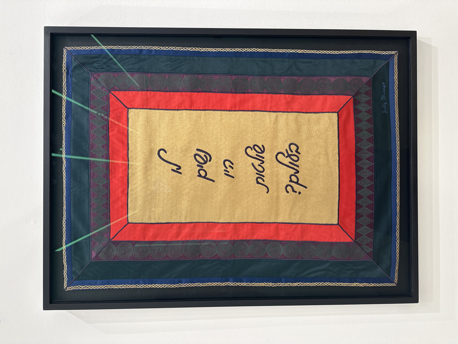 A framed textile artwork hangs centered on a plain white wall in a brightly lit interior space, indicative of a gallery or exhibition. The artwork is rectangular, oriented vertically, and encased in a simple black frame.
The textile piece features a central gold-colored fabric panel with five lines of black Hebrew script. This central panel is bordered by multiple layers of fabric. Immediately surrounding the gold panel is a solid red fabric border. Outside the red border, there is a dark green or deep teal fabric border. This dark green/teal border displays a repeating pattern of dark gray/purple diamonds along its top and bottom horizontal segments, and a repeating spiral pattern along its vertical segments. Emanating upwards from the top edge of the central gold panel, extending into the upper dark green/teal border, are several bright green, thin, luminous lines. The outermost fabric border is a dark blue, accented with a thin, light-colored (likely gold or beige) braided trim. In the lower right corner of the dark green/teal border, the signature "Judy Chicago" is visible in black cursive script.
The specific location within London, United Kingdom, is not discernible from the provided image.