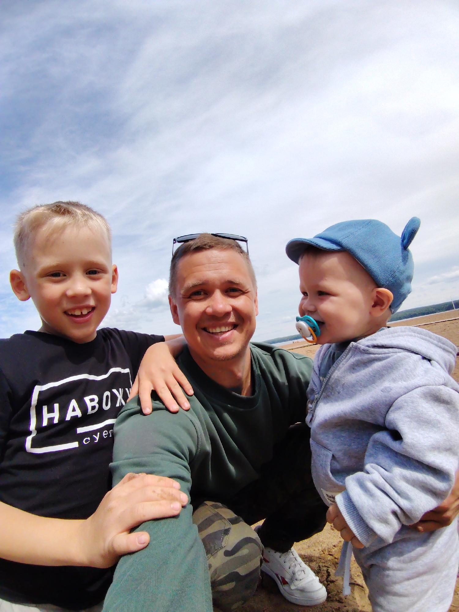 A joyful family portrait features three males—an adult and two young boys—captured in a close-up, slightly wide-angle outdoor selfie. The adult, positioned centrally, is smiling broadly with short dark hair and sunglasses perched on his head. He wears a dark green long-sleeved shirt, camouflage pants, and white sneakers with red accents.
To his left, an older boy with blond hair also smiles widely, with his arm resting on the adult's shoulder. He is dressed in a black short-sleeved t-shirt featuring white block text that appears to read "HABOX" and smaller text below, possibly "CYET". To the adult's right, a toddler with a blue cap, which has ear-like protrusions, looks to the side with a slight smile. He has a blue, white, and orange pacifier in his mouth and wears a light grey hooded sweatshirt.
The scene takes place outdoors on a bright day, under a clear sky with scattered white clouds. The ground is sandy or dusty, consistent with a riverbank or open area, aligning with the location context of Chaikovsky, Russia, which is on the Kama River. A faint line of distant land or trees is visible in the background. All individuals exhibit happy expressions, suggesting a cheerful family outing.