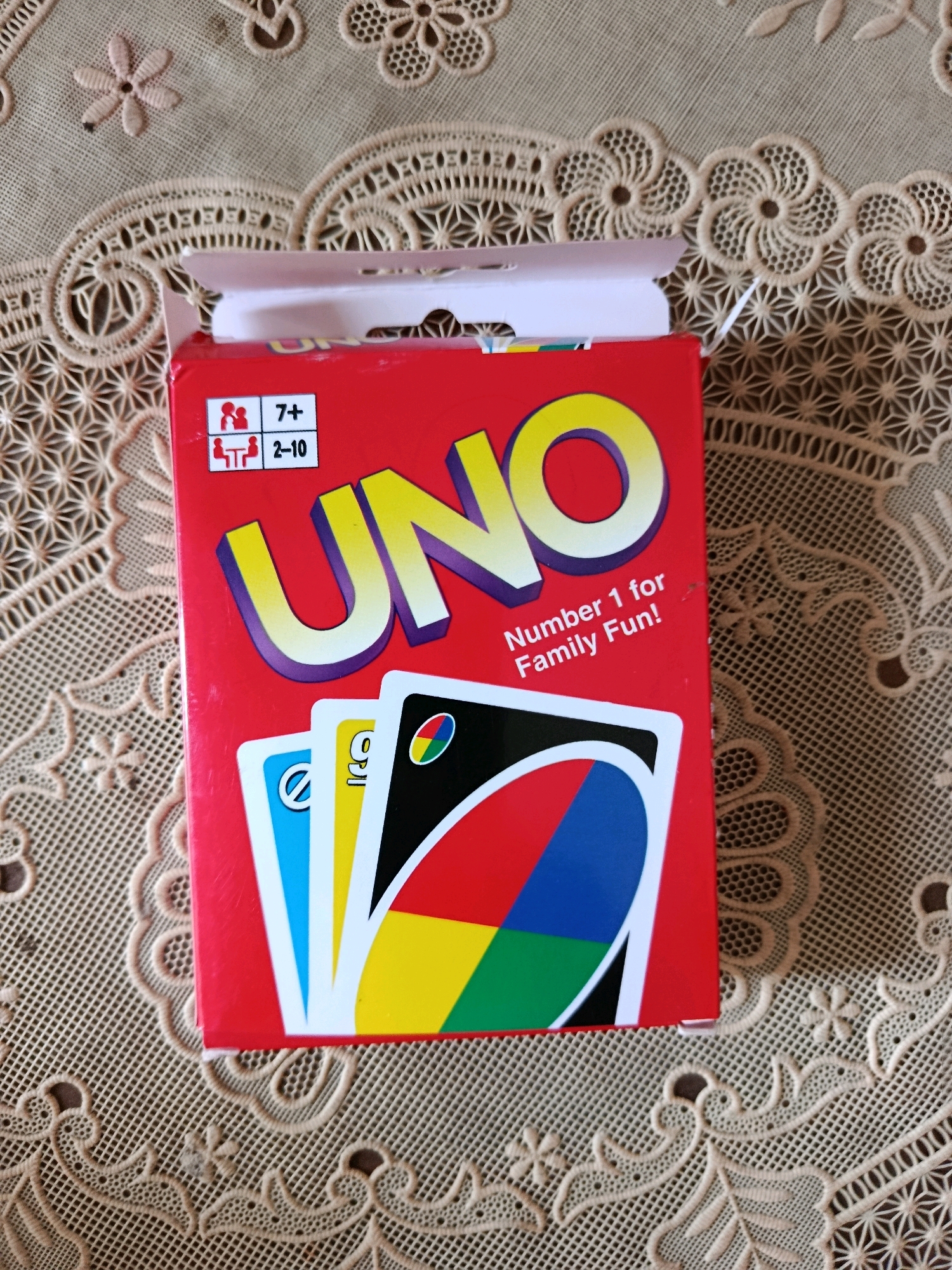 The image shows a red UNO card game box placed on a beige, lacy tablecloth. The box is slightly ajar, with its lid partially open. The UNO logo, in large yellow letters with purple outlines, is prominently displayed on the front of the box. Below the logo, the text "Number 1 for Family Fun!" is visible. To the left of the logo, there are icons indicating age and player count: a silhouette of people and the number "7+" for age, and two silhouettes of people at a table with the number "2-10" for player count. A few UNO cards, showing parts of their designs (blue, yellow, and a black card with a colored circle), are peeking out from the top of the box. The tablecloth has intricate floral and geometric patterns. The scene appears to be indoors, possibly on a table. There are no people visible in the image, and no specific identifiable landmarks to place it in Jambearum, Indonesia.