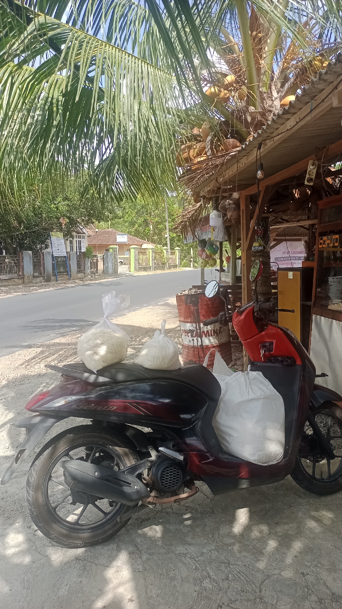 The image depicts a roadside scene in Cibeber, Indonesia. In the foreground, a dark red and black scooter is parked on a dusty, paved surface. Two large, translucent plastic bags filled with a white, granular substance, likely rice or similar foodstuff, are placed on the scooter's seat and rear. Next to the scooter, a red and white oil drum with the word "PERTAMINA" printed on it is visible. Behind the drum, a small roadside stall with a corrugated metal roof made of palm fronds is set up. Coconuts hang from a palm tree above the stall. The stall appears to be selling various items, including what look like small toys or decorative objects hanging from the roof. A sign with Indonesian text is affixed to the stall. In the background, a road stretches into the distance, flanked by trees and houses with tiled roofs. The sky is blue and partially obscured by the fronds of a palm tree.