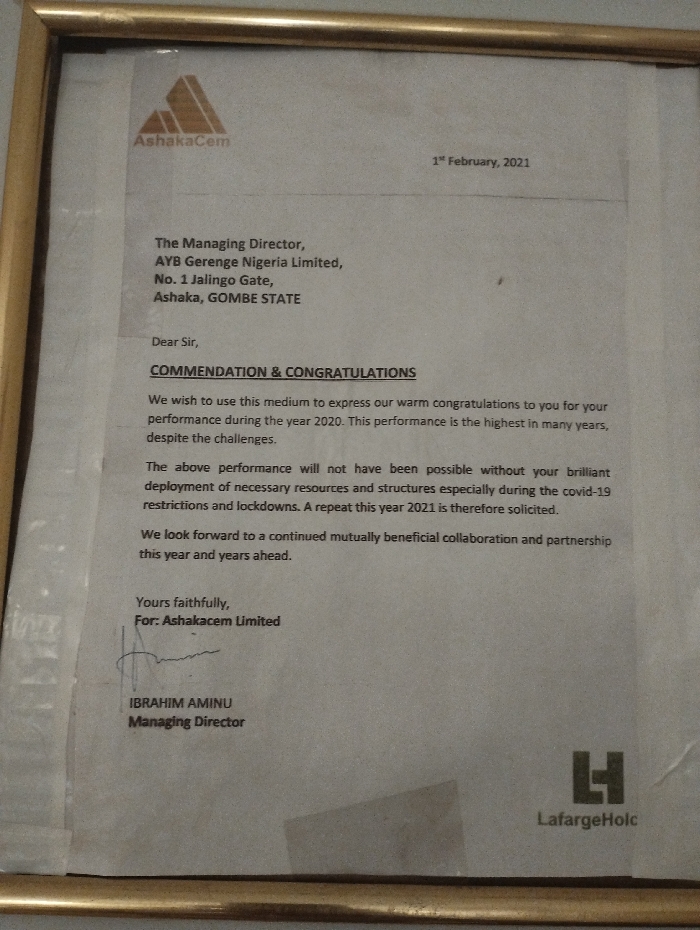 The image is a framed letter on a white background. The letter is from AshakaCem to an individual. The letter is dated 1st February 2021. It is addressed to Dear Sir, and expresses commendation and congratulations for the individual's performance in the year 2020. The performance is described as the highest in many years, despite the challenges. The letter goes on to say that the performance would not have been possible without the individual's brilliant deployment of necessary resources and structures especially during the COVID-19 restrictions and lockdowns. The letter ends by saying that they look forward to a continued mutually beneficial collaboration and partnership this year and years ahead. The letter is signed Yours faithfully, For: Ashakasem Limited. The signature reads Ibrahim Aminu, Managing Director. The letterhead has the logo of AshakaCem and a smaller logo of LafargeHolc. The coordinates provided (latitude: 10.917, longitude: 11.476) correspond to the city of Ashaka, Nigeria, where AshakaCem is located. There are no other people present in the scene, and no other objects or actions can be determined from the image.