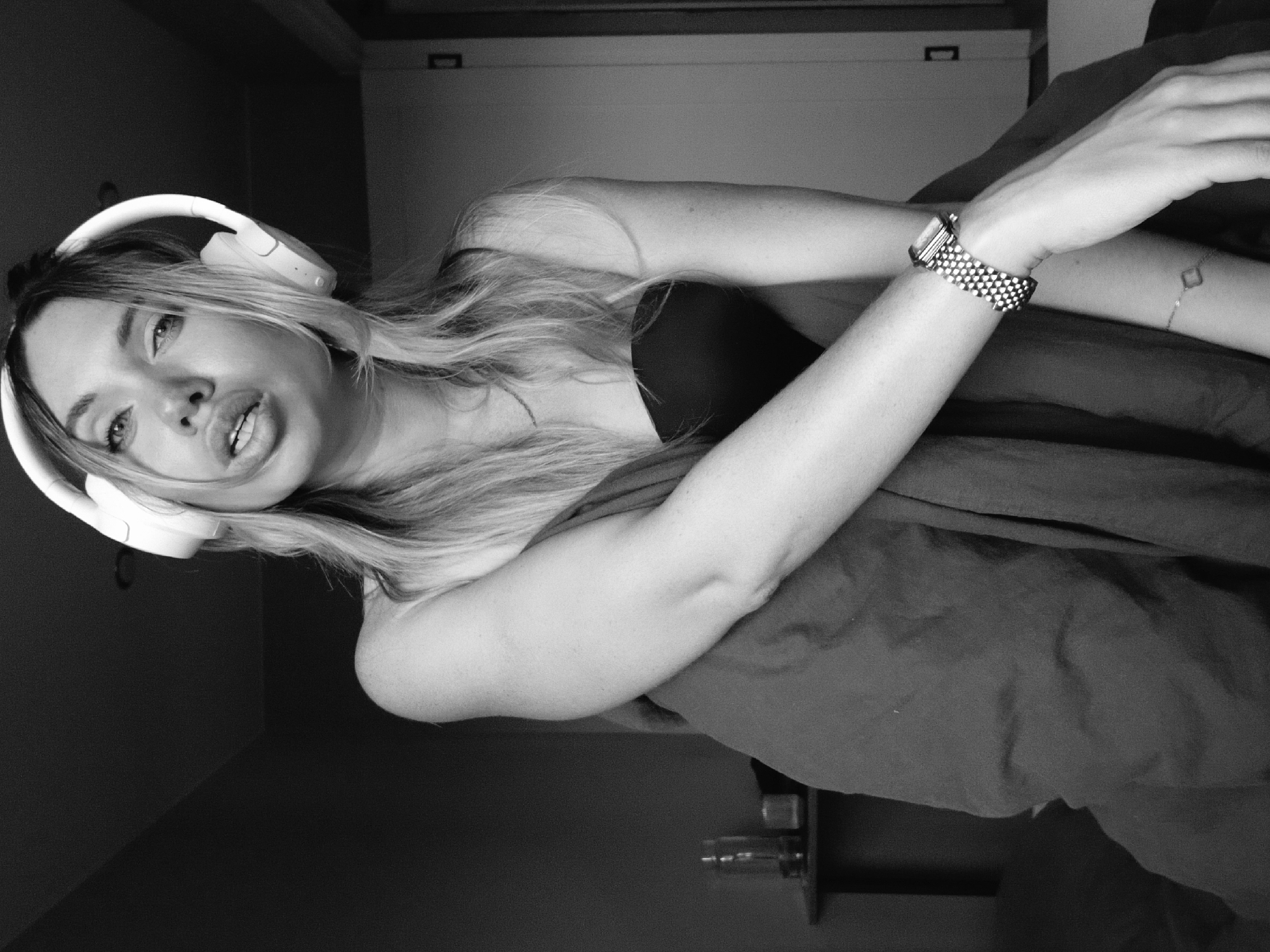A woman with long, light-colored hair is wearing white headphones and resting on what appears to be a bed covered with dark bedding. She is wearing a watch and a bracelet on her wrist. The photograph is in black and white. The background includes a plain white door or wardrobe and a dark corner. The location in Votkinsk, Russia, cannot be determined from the visual information provided.