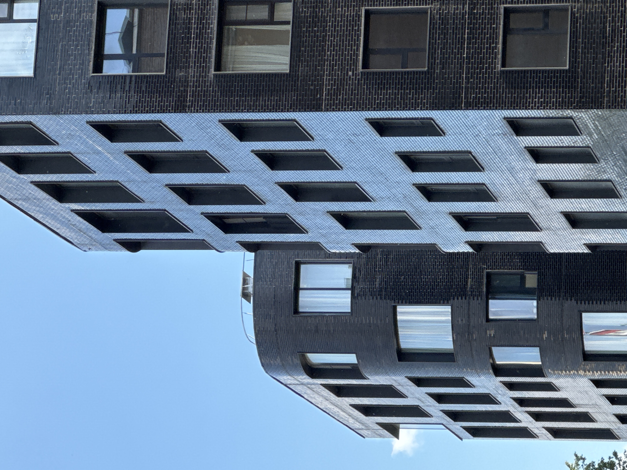 The image captures a modern building in Moscow, Russia, against a clear blue sky. The photograph is oriented sideways, presenting the building horizontally across the frame.
The building's structure features a dark, possibly black or very dark grey, textured facade, resembling brickwork, particularly in the upper section of the visible architecture. This primary facade is punctuated by rectangular windows with dark frames, which reflect the bright sky and sometimes internal light.
Below this main section, a distinctive architectural element extends, appearing as a patterned overlay or an integrated lower part of the building. This element creates a grid of dark, recessed rectangular openings, arranged in consistent rows. The surface of this patterned section exhibits a lighter tone, potentially due to reflections or its material, and has a subtle diagonal striped texture, occasionally reflecting the blue of the sky. One edge of the building, curving towards the bottom right of the image, includes a visible railing, suggesting a roof terrace or balcony area.
The scene is bathed in bright daylight under a clear sky, with a small white cloud visible towards the lower edge of the frame. No people or visible text are present in the image. The overall impression is of contemporary urban architecture, focusing on geometric patterns, contrasting textures, and reflective surfaces.