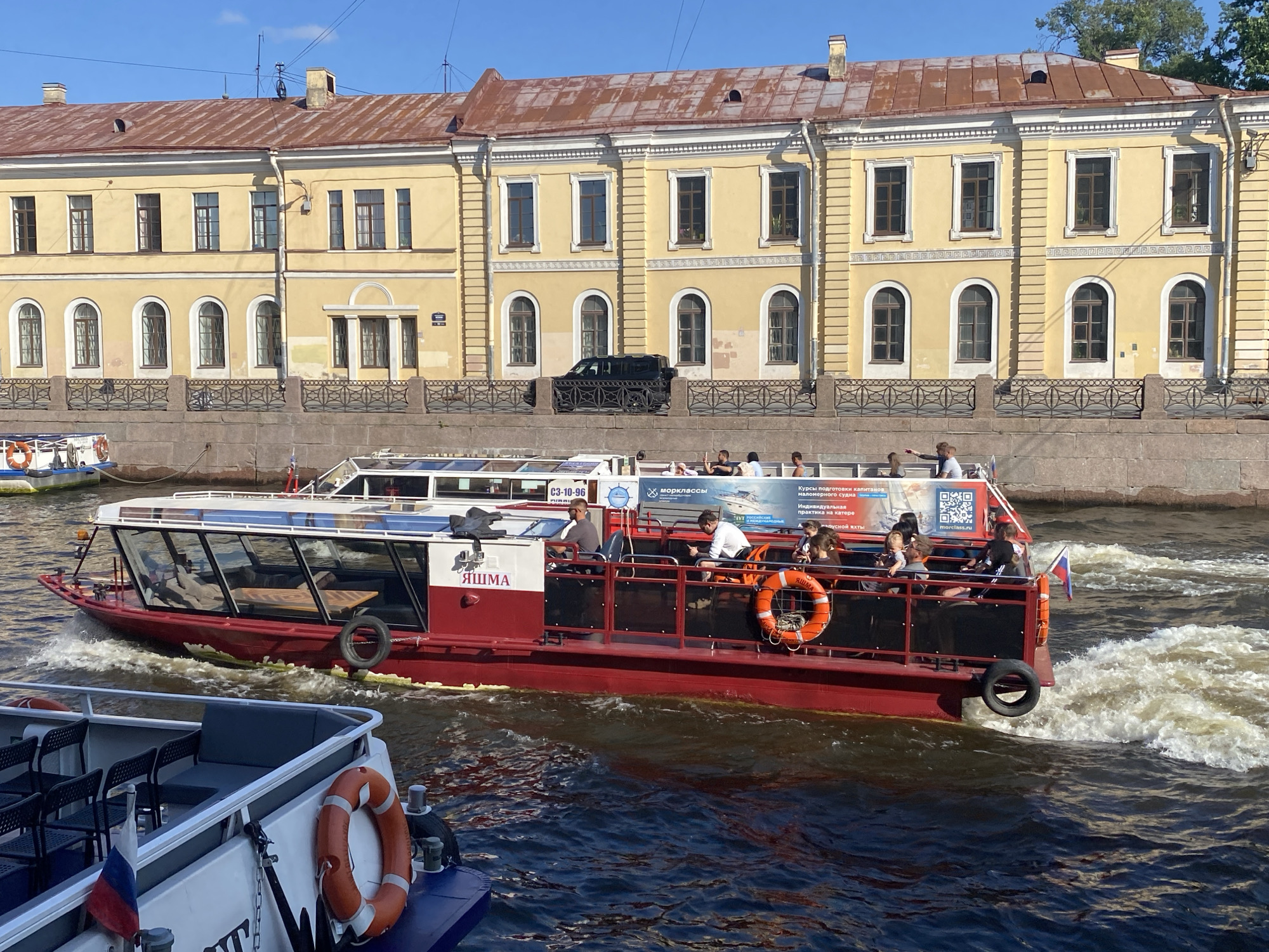 The image shows a bright, sunny day in Saint Petersburg, Russia, with a canal in the foreground and a historic building in the background. A red and white tour boat is navigating the canal, creating white foam in its wake. Several passengers are visible on the boat. On the lower left of the image, a portion of another boat is visible, also with seating and a life preserver. The building in the background is a long, yellow, three-story structure with a rust-colored roof. It has numerous windows and arched doorways, indicative of historical architecture. A black SUV is parked in front of the building. The water of the canal is dark blue, with the sunlight reflecting off its surface.
