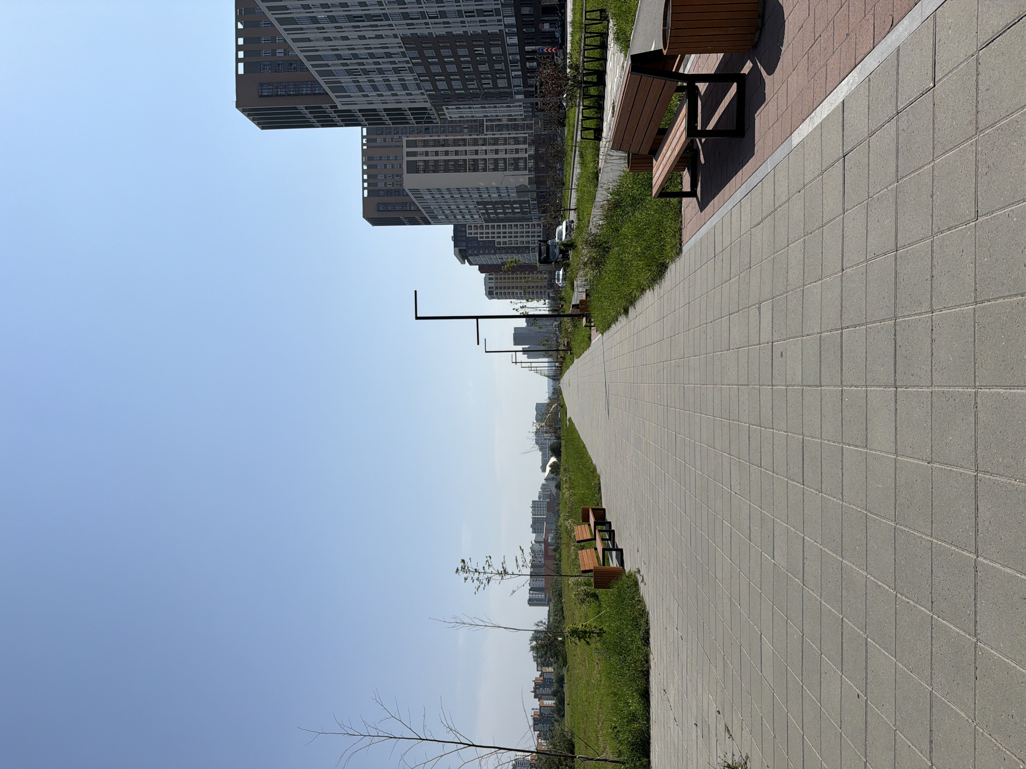 The image depicts a modern urban park pathway with a clear blue sky overhead. The pathway is paved with large, light-colored square tiles and runs parallel to a green grassy area on the left and a road with buildings on the right. Several wooden benches are placed along the pathway, some with integrated trash bins. Tall, contemporary residential buildings with numerous windows dominate the right side of the scene, suggesting a developed urban area. In the distance, a cityscape with more buildings is visible, along with sparse trees. The overall impression is one of a new, well-maintained public space designed for recreation and urban living. Based on the architecture and the presence of modern infrastructure, this scene is likely located in a newly developed district of Yekaterinburg, Russia, such as the Akademichesky district, known for its contemporary residential complexes and planned green spaces. There are no people visible in the image, and no specific actions or interactions are taking place.