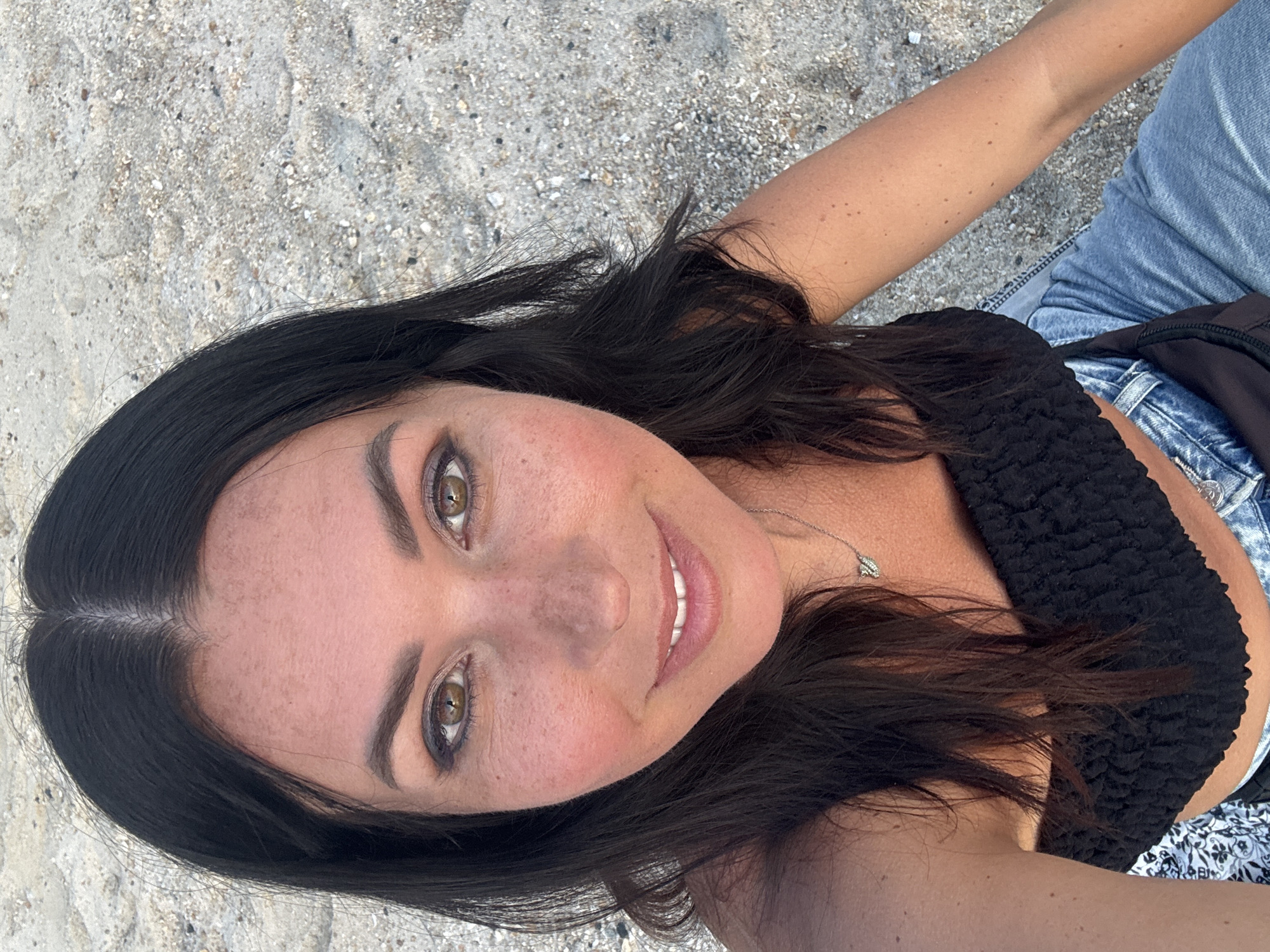 A woman is posing outdoors, likely on a beach, given the sandy ground in the background. She is wearing a black textured top and light blue denim jeans. She has long, dark hair and fair skin with freckles. She is smiling, and the image is taken at a slightly upward angle. The location is possibly Baan Sri Thanu, Thailand.