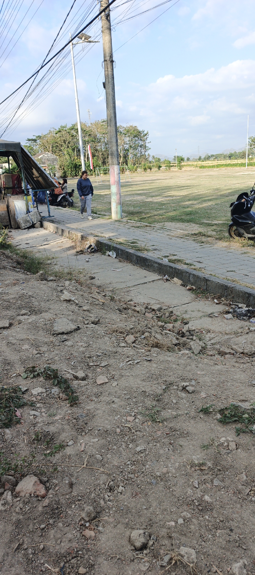 A woman in a blue sweatshirt and light grey pants walks on a paved sidewalk in Kasiyan Timur, Indonesia. She is facing left and appears to be walking towards the left side of the image. To the left of the sidewalk, there is a small open-air stall with a dark-colored roof, and motorcycles are parked behind it. The sidewalk is bordered by a rough, dirt area on the left with scattered debris, and a grassy field on the right. A utility pole with multiple wires is visible behind the woman. The sky is partly cloudy.