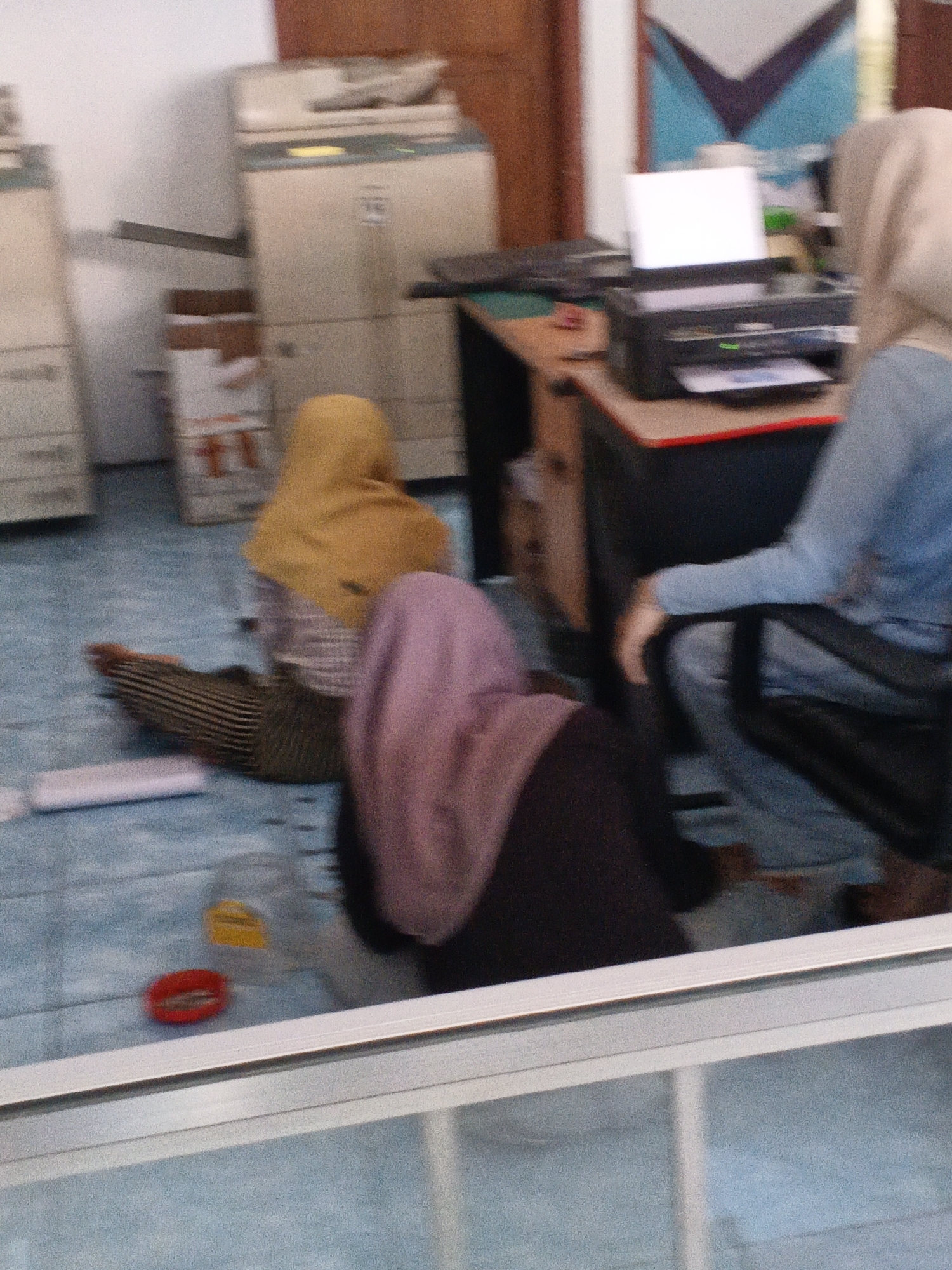The image depicts an indoor scene with several individuals, possibly in an office or a communal space. The floor appears to be tiled in a light blue color, suggesting a clean and functional environment. In the background, there are office machines, including a photocopier, and stacked boxes, indicating a work or storage area. Three individuals are partially visible, each wearing a hijab, which is common attire for women in Indonesia. One person is seated at a desk with a laptop and printer. Another individual is lying on the floor, partially covered by a striped blanket, suggesting they might be resting or sleeping. A third person is also on the floor, wearing a dark purple hijab and outfit. The overall impression is one of a casual and perhaps somewhat informal setting within an Indonesian office. The presence of the specific office equipment, the tiled floor, and the attire of the individuals strongly suggest this scene is located in Padang, Indonesia.