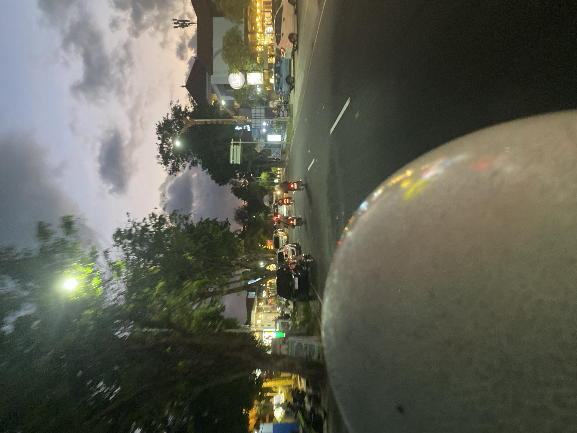 The image shows a street scene in Kuta Selatan, Indonesia, at dusk. The view is from a vehicle, possibly a motorcycle, with part of the helmet visible in the lower right corner. A two-lane road runs into the distance, with multiple motorcycles and a black car on it. Buildings with lit windows are present along the street, and trees are lining the roadside. Streetlights illuminate the scene. The sky is cloudy with colors of dusk.