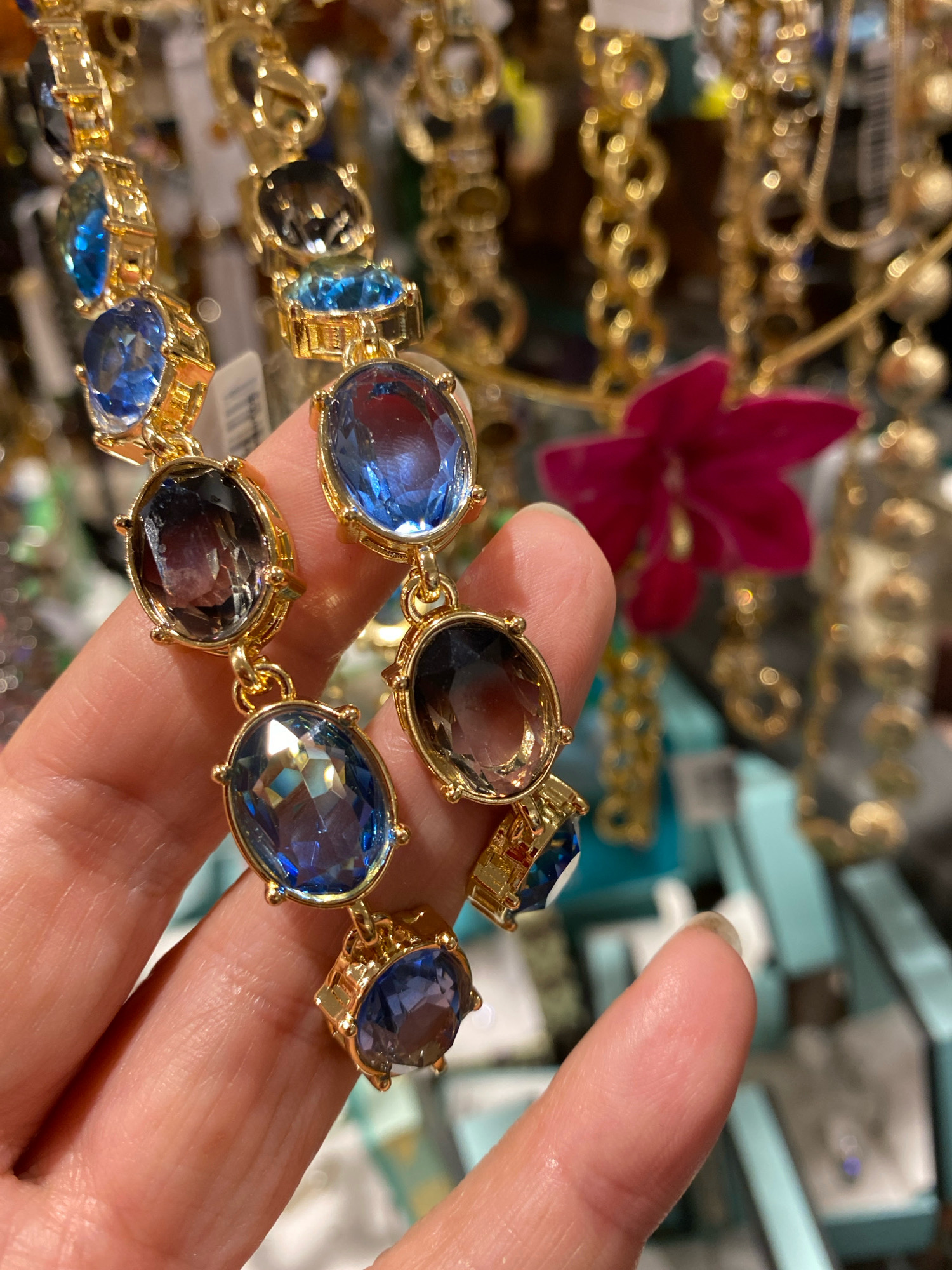 A hand holds a gold necklace with oval-shaped blue and smoky quartz gemstones. The necklace is made up of multiple dangling charms, each featuring a large oval gemstone set in gold. The gemstones vary in color from deep blue to lighter blue and some are smoky or greyish. The background is blurred, showing other jewelry displays, a bright pink flower, and hints of turquoise boxes, suggesting a retail environment such as a jewelry store. The image is taken in a close-up perspective, focusing on the necklace and the hand holding it.