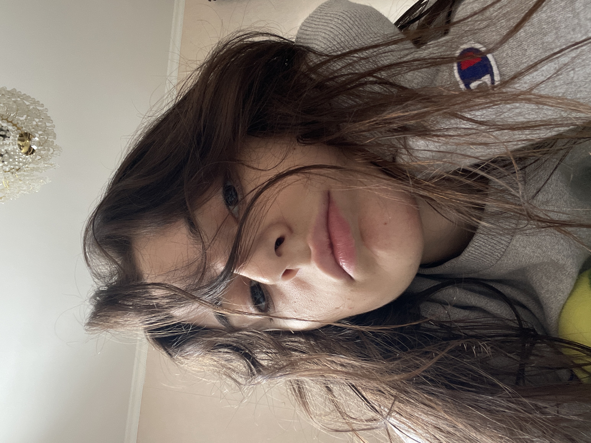 An close-up, rotated portrait shot captures a person with long, dark brown hair flowing around their face. The image is oriented with the subject's head tilted significantly, making it appear as if they are lying down or captured from a unique, low-angle perspective.
The subject's face is central, with one eye visible and dark in color, looking towards the viewer. Their nose and slightly parted lips, which appear naturally pink and glossy, are prominent. Strands of their rich brown hair fall across their forehead and cheeks, adding to a casual, perhaps contemplative, look.
The person is wearing a grey sweatshirt, and a distinctive "Champion" logo, featuring blue, red, and white, is clearly visible on the upper left chest area.
The background primarily consists of a light-colored ceiling on the left, where a ornate, possibly crystal or beaded, chandelier is partially visible. On the right, a bright yellow or lime green object, possibly a cushion or garment, can be faintly seen. The overall setting suggests an indoor environment, likely a home in Moscow, Russia, bathed in soft, diffused light. The expression of the subject is neutral, bordering on pensive.