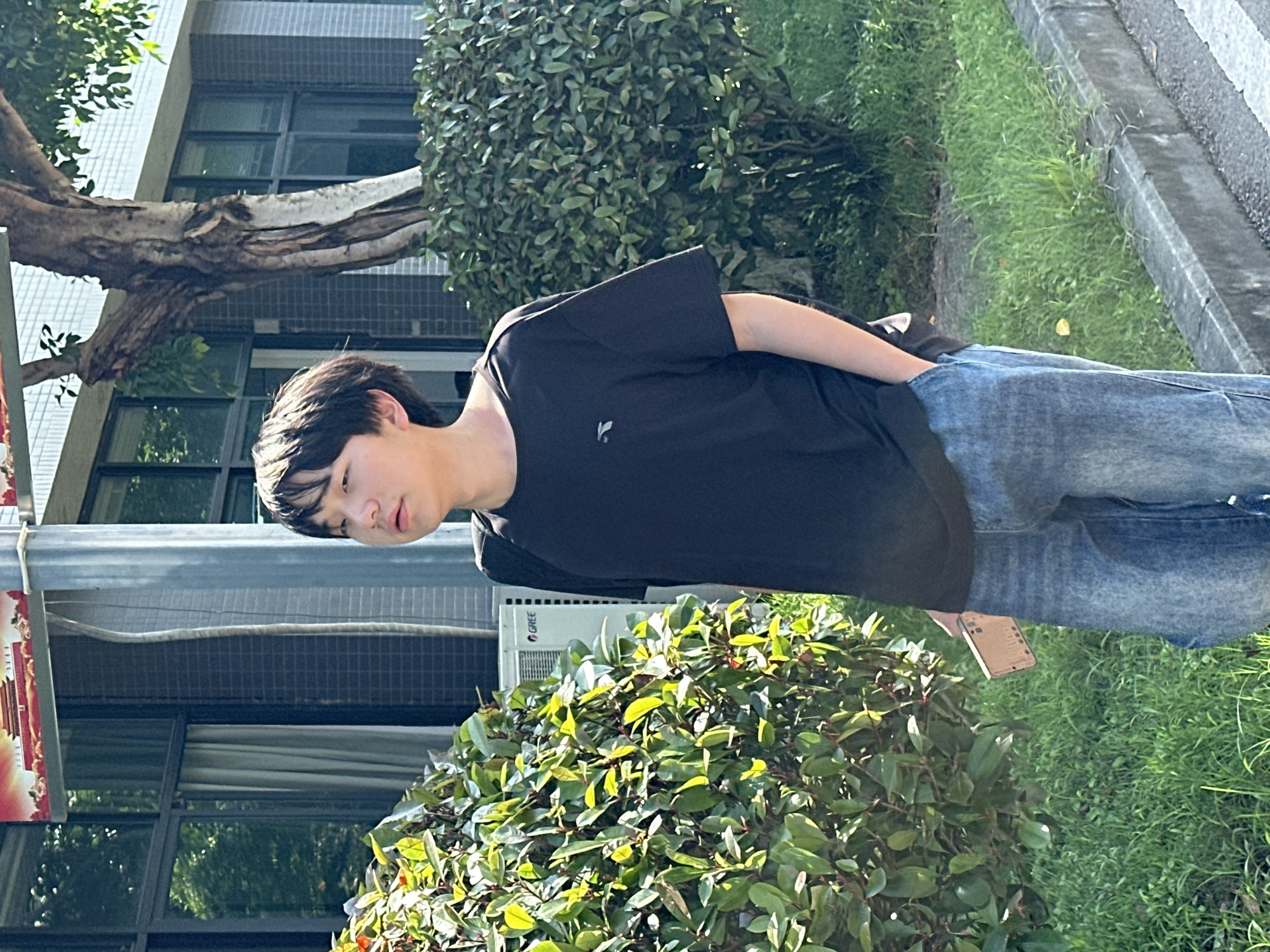 This outdoor photograph, taken in bright daylight in Jiangjin, China, features a young man standing casually amidst greenery and urban architecture.
The subject is a young man with short dark hair, dressed in a plain black short-sleeved t-shirt and blue denim jeans. He stands with his right hand in his jeans pocket, looking downwards and slightly to his left with a neutral or slightly pensive expression. A light-colored smartphone with a brown or tan case is visible, partially tucked into his back right pocket. A small, white, stylized logo, possibly depicting a bird or abstract design, is visible on the left chest of his t-shirt.
The background shows a building with a textured, light-colored facade and dark-framed windows. A large tree with a thick, gnarled trunk and branches extends horizontally from the left side of the frame, partially obscuring the building. The foreground and midground are dominated by dense green bushes and shrubbery. Behind one of these bushes, an outdoor air conditioning unit with the brand "GREE" clearly visible on its side is mounted on the building. To the right, a strip of green grass runs alongside a concrete curb. A red and white sign or banner, possibly depicting a tiered building or temple, is partially visible on the far left, attached to a pole.
The scene appears to be a relaxed, everyday moment under clear, bright weather conditions.