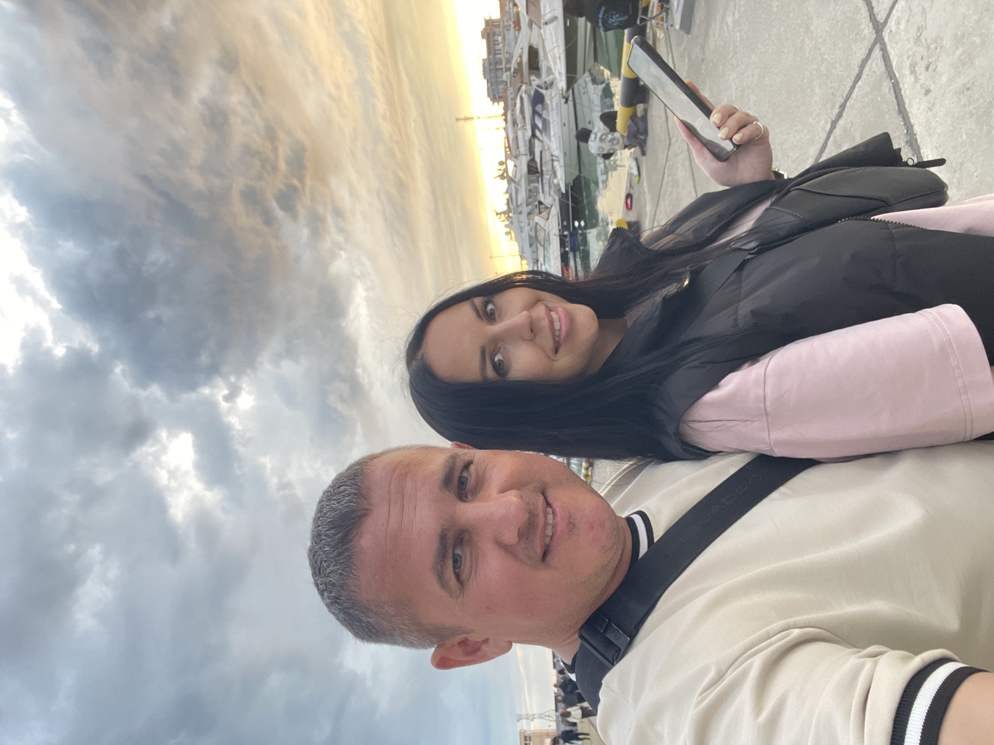 Two people are posing for a selfie against the backdrop of a marina. The marina is located in Sochi, Russia. A woman with dark hair holds a smartphone. The man has short, gray hair. The background shows docked boats, buildings, and a sky with a mix of clouds and the warm light of dusk or dawn.