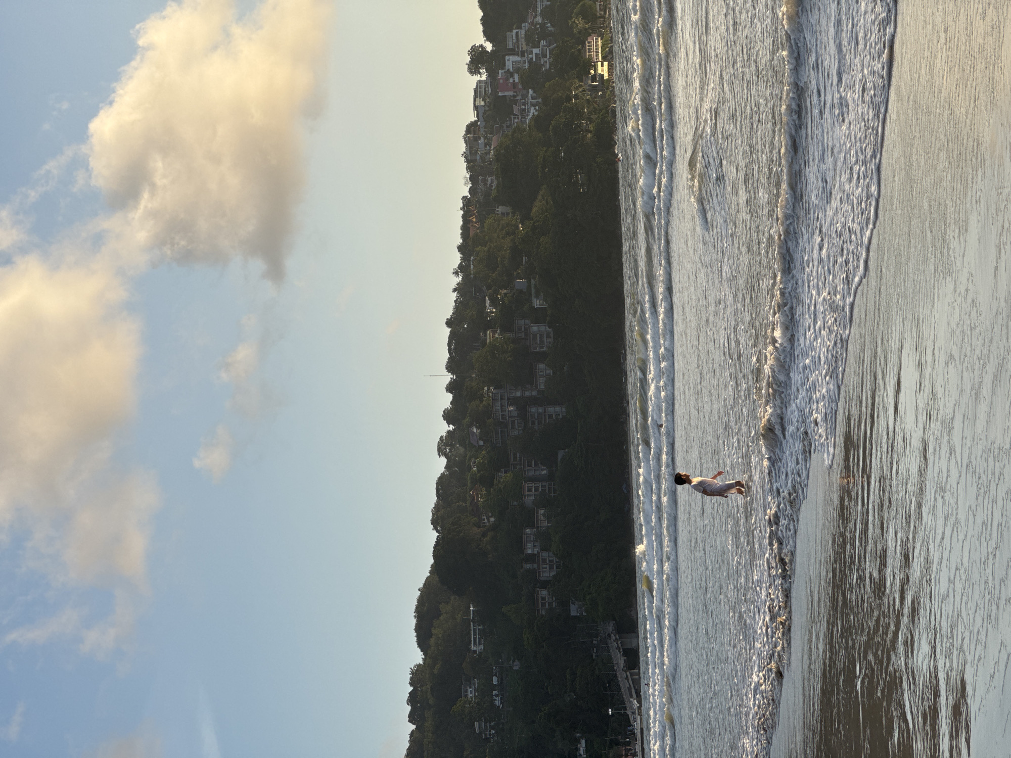 A lone child, wearing a white outfit, walks through shallow ocean water near the shore of Patong Beach in Thailand. The water has gentle waves and white foam. In the background, a lush, green hillside is covered with numerous buildings, including houses and hotels, interspersed with trees. The sky above is a pale blue with scattered, wispy clouds, some of which are tinged with yellow light from the setting sun. The overall scene depicts a tranquil beach environment with a backdrop of a developed coastal hillside.