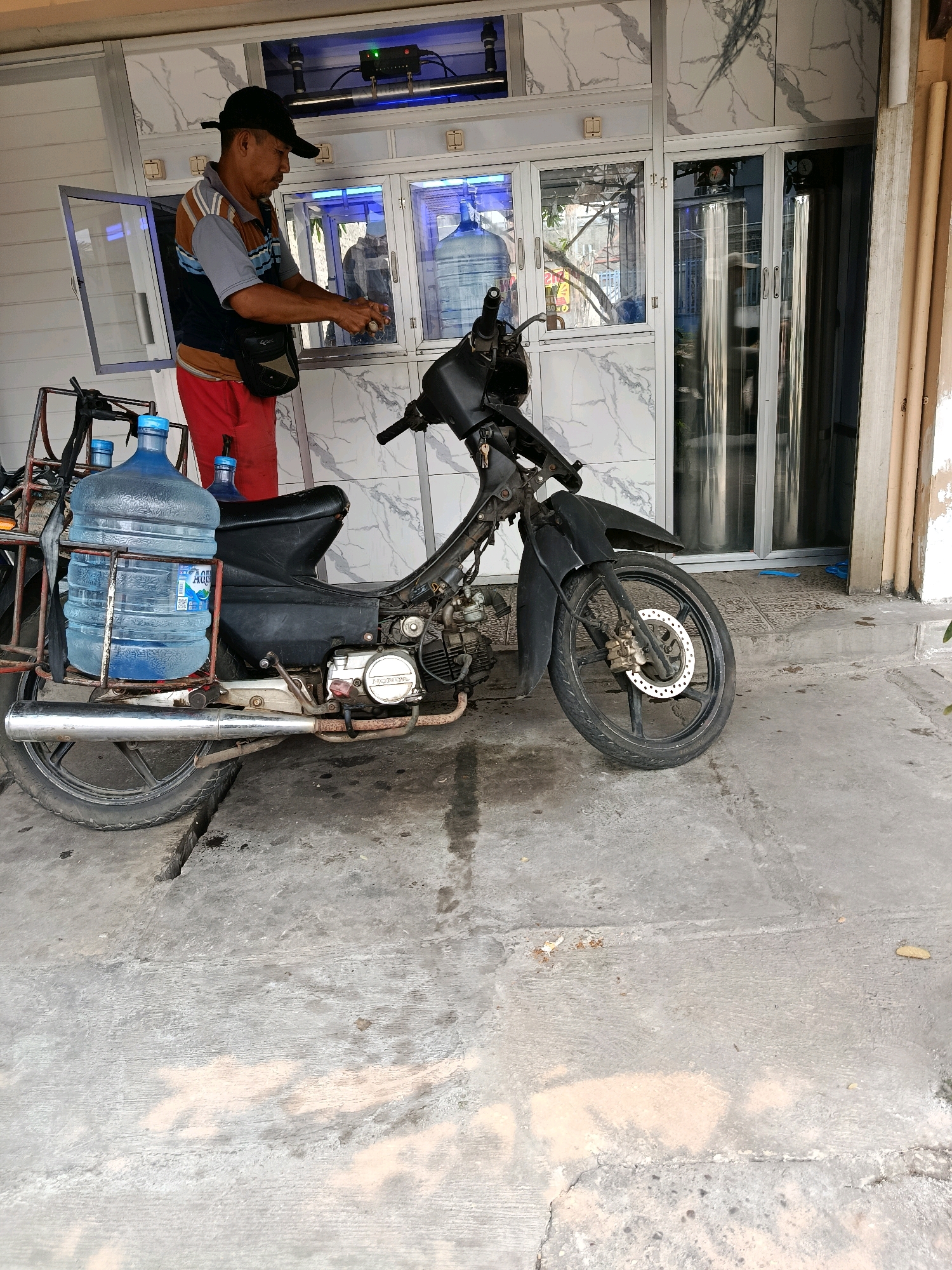 In Bekasi, Indonesia, a man wearing a black cap, a grey and brown striped shirt, and red pants is standing next to a black motorcycle. The motorcycle has two large blue water jugs strapped to a rack at the back. The man appears to be in the process of handling or dispensing something, with his hands held out in front of him. Behind him is a building with a white marble-patterned facade and glass-fronted compartments containing more water jugs. The scene is set on a concrete surface, possibly a sidewalk or street.